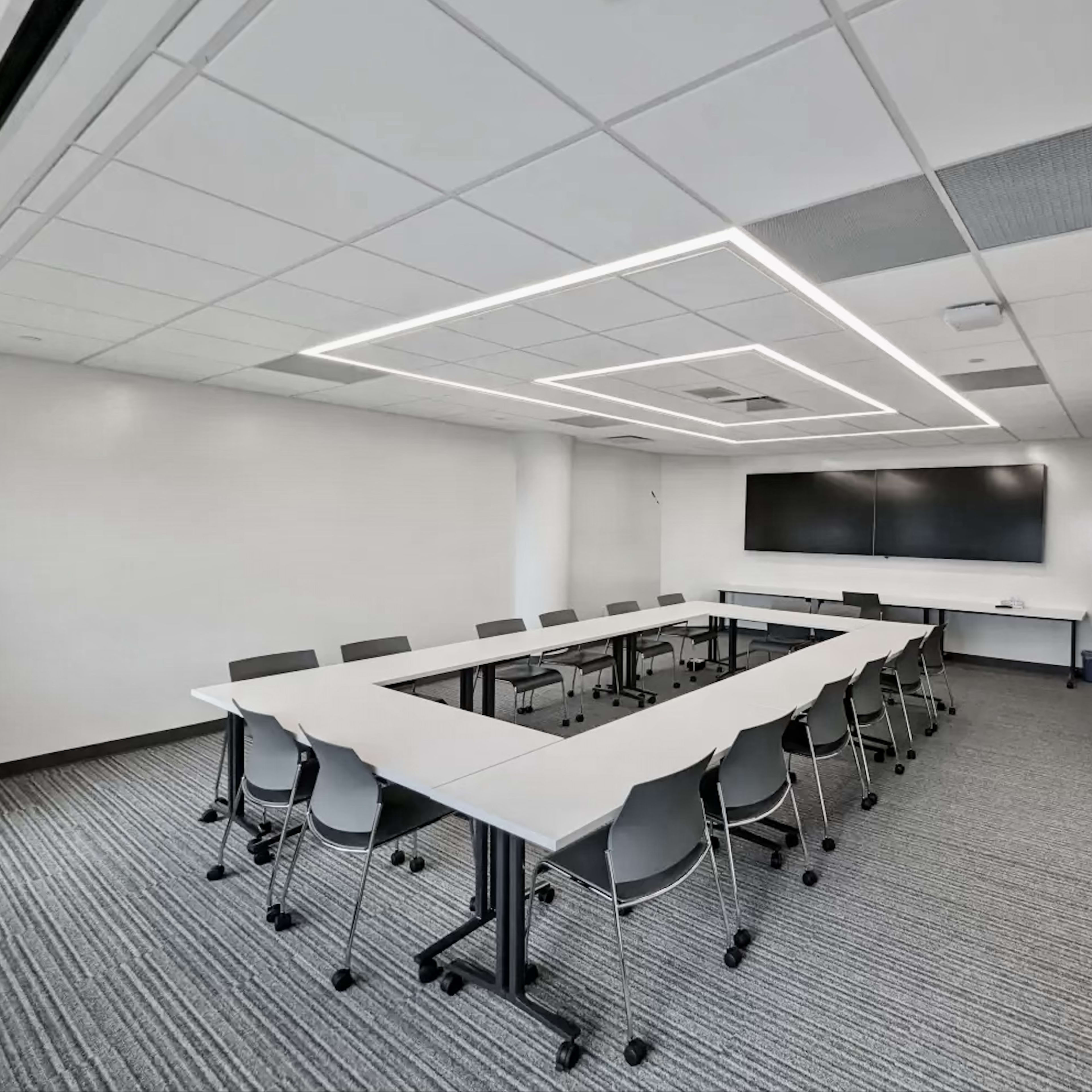 A conference room features a rectangular table surrounded by chairs, with a large screen mounted on the wall.