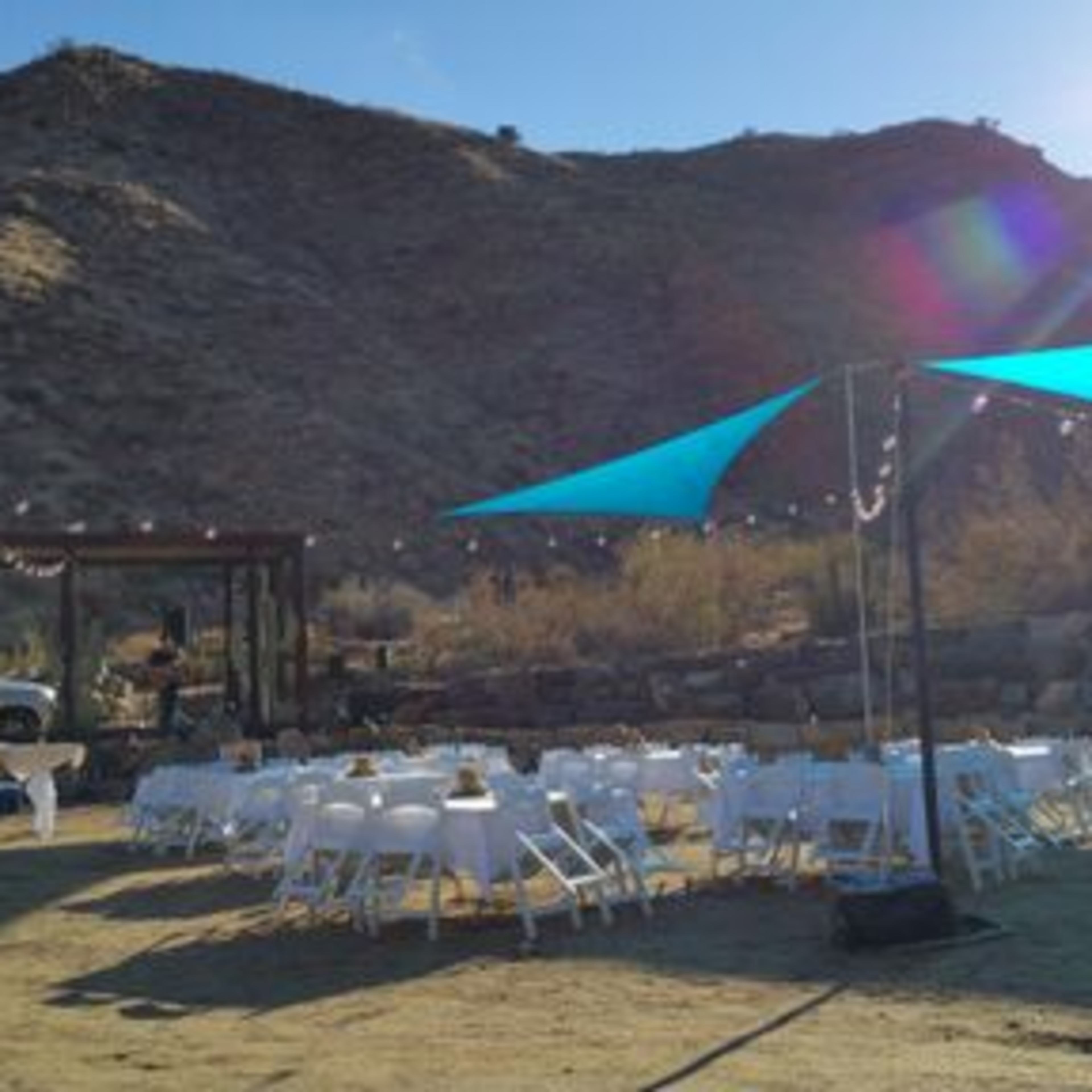 A landscape setup for an outdoor event features several tables with white tablecloths under blue shade sails, surrounded by hills.