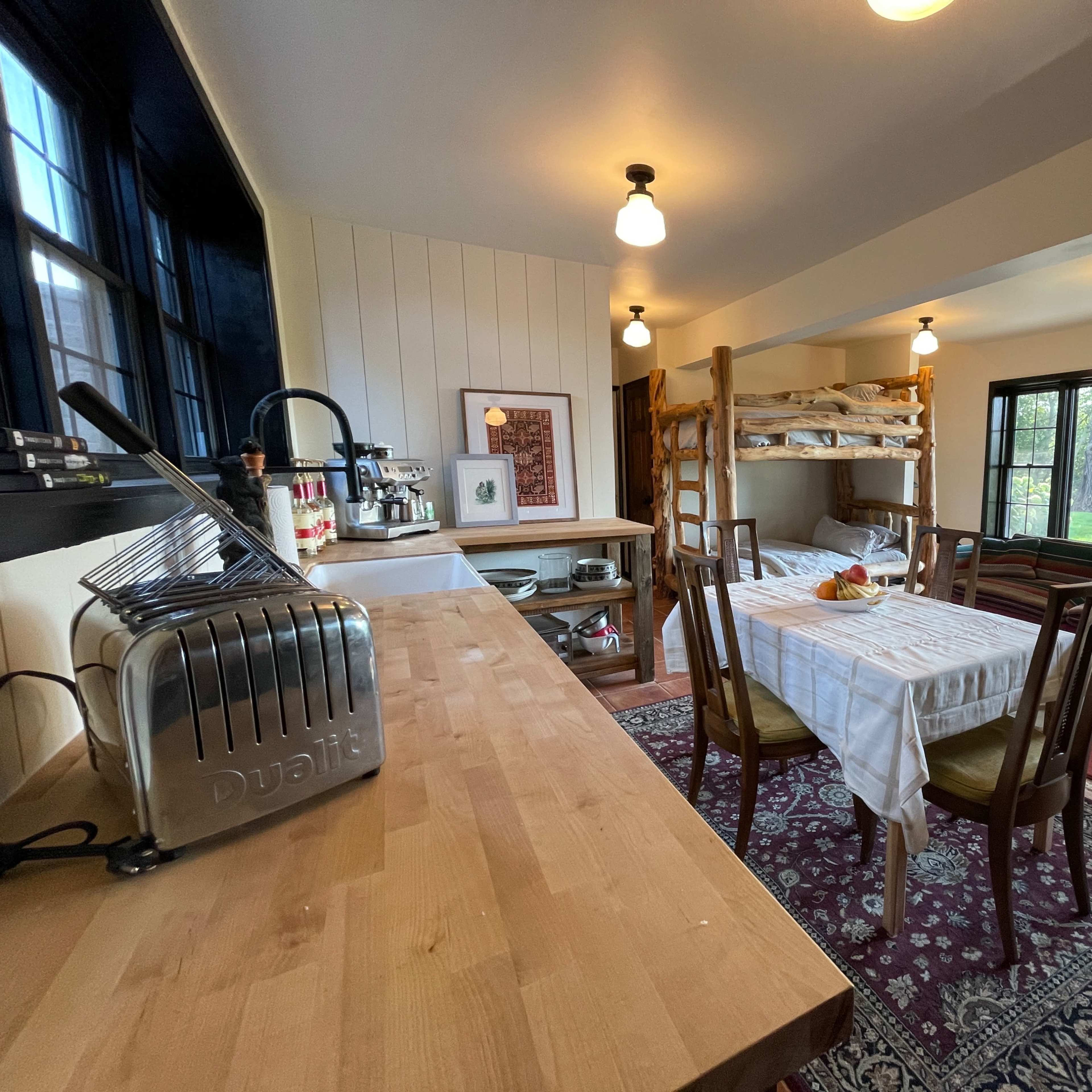 A cozy, open-concept living space featuring a kitchen area with a wooden table, a small dining area, and a bunk bed beside large windows that provide a view of the outdoors.