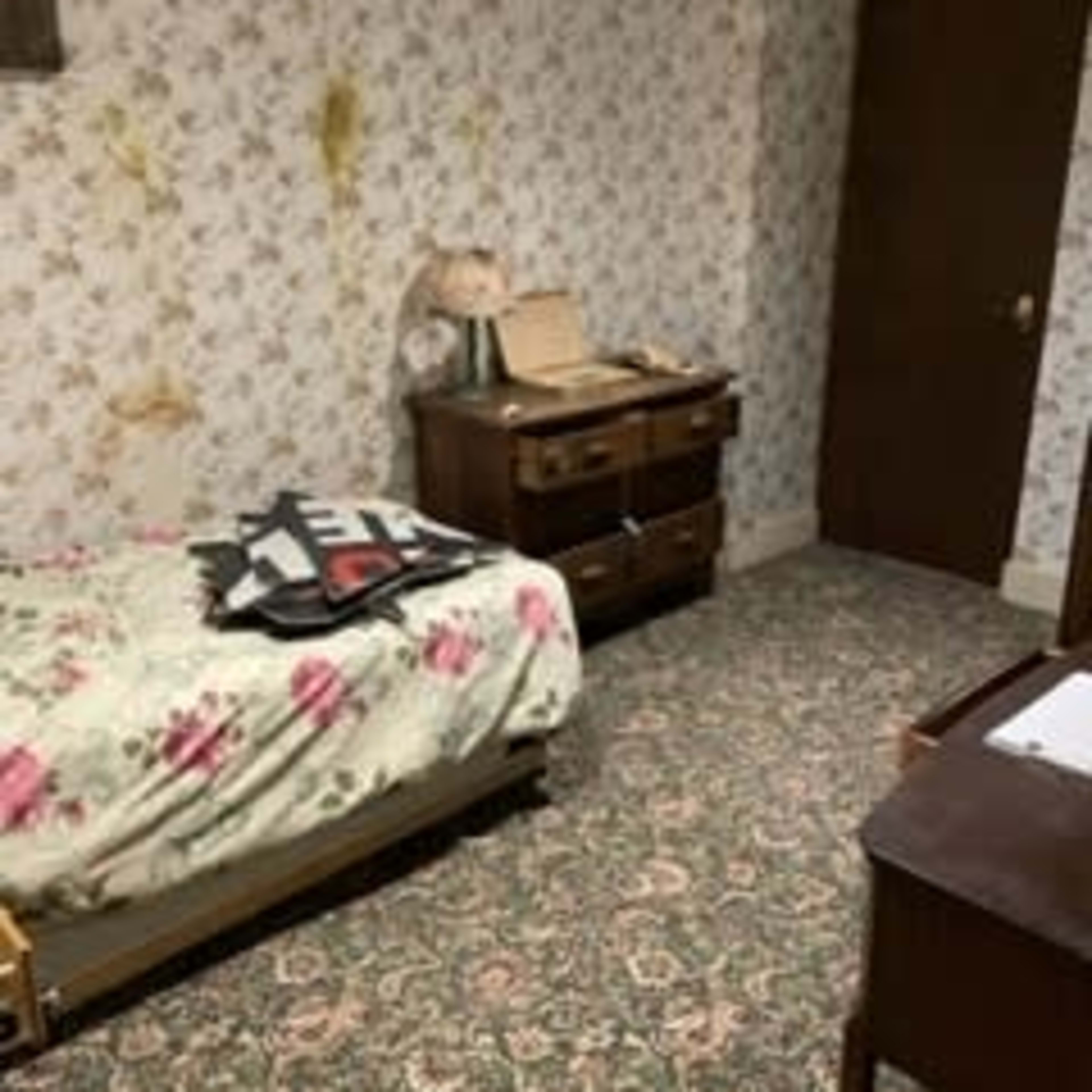 The room features a floral-patterned bedspread on a bed, a wooden dresser, a lamp, and a patterned carpet against wallpaper adorned with a floral design.