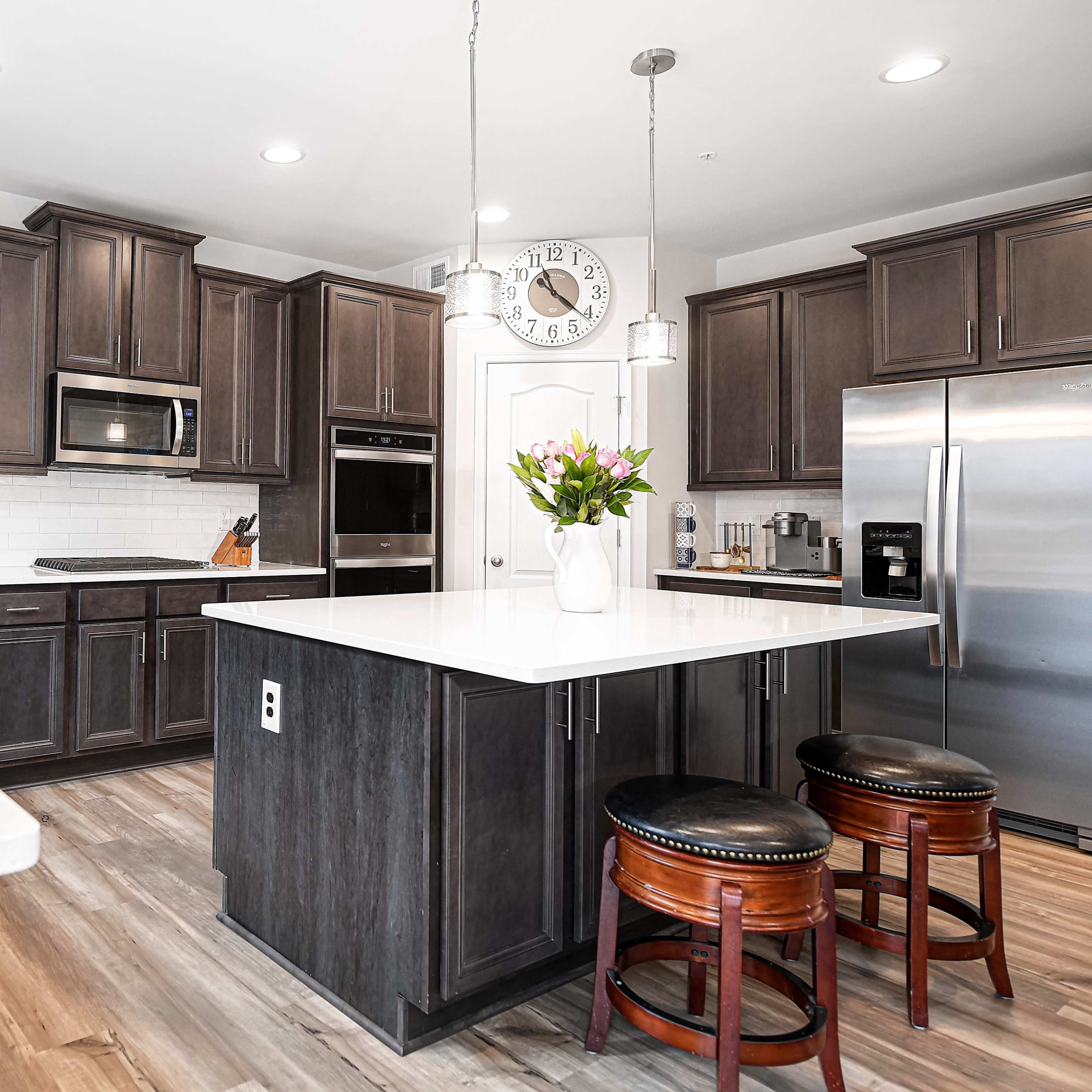 A modern kitchen with dark wood cabinets, stainless steel appliances, a large island with white countertops, and a flower vase on the counter.