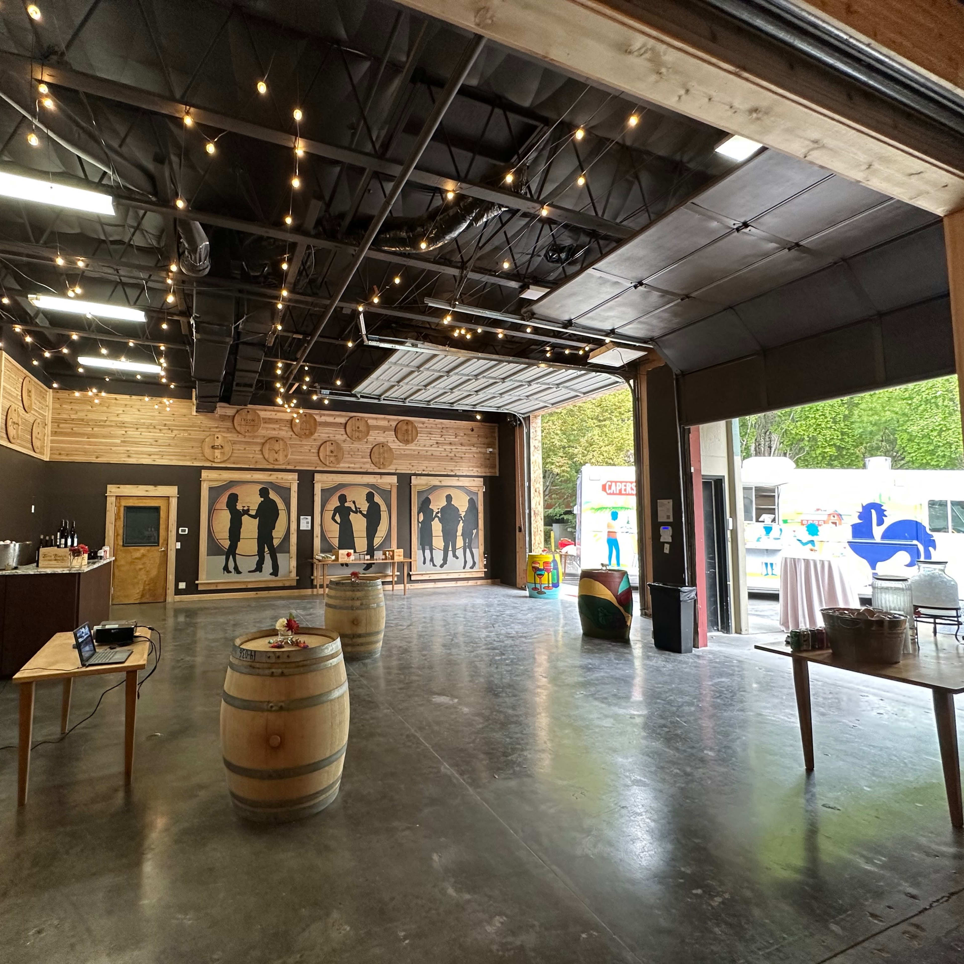 An indoor event space features wooden walls, silhouetted figures, wine barrels, and string lights.