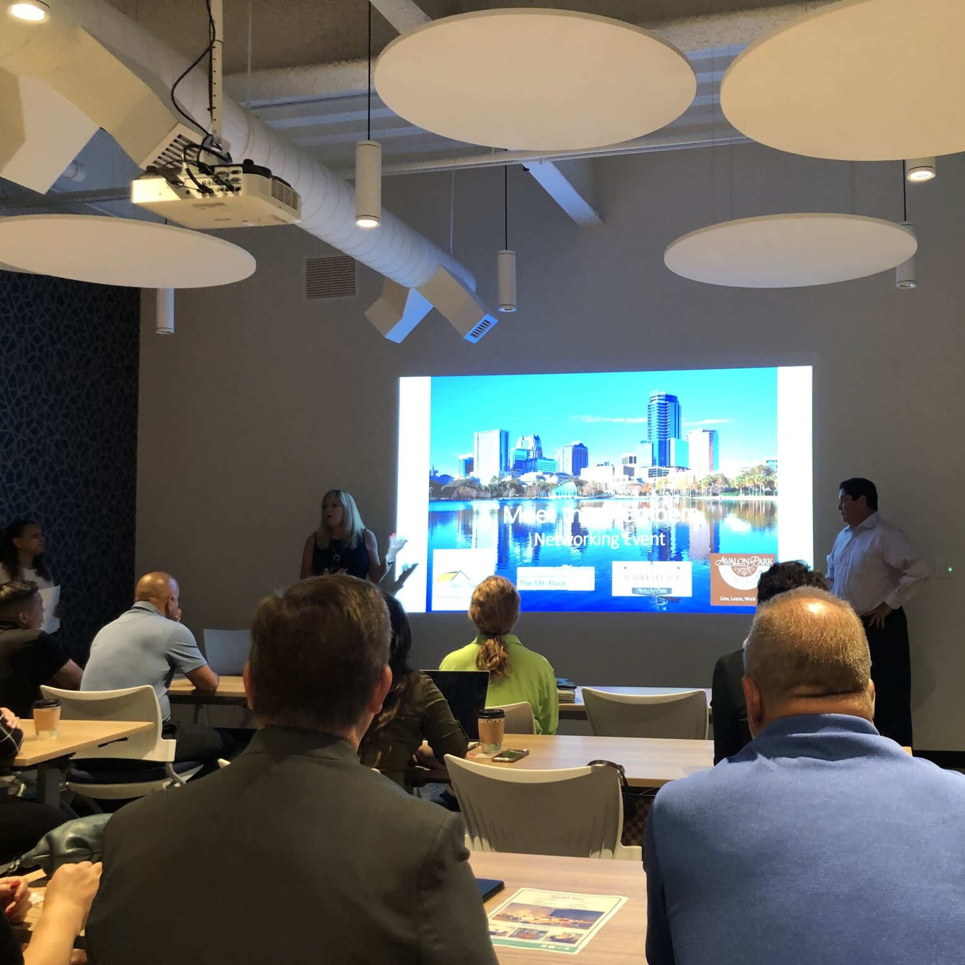 A group of people sits in a conference room, watching a presentation on a large screen that displays a city skyline.