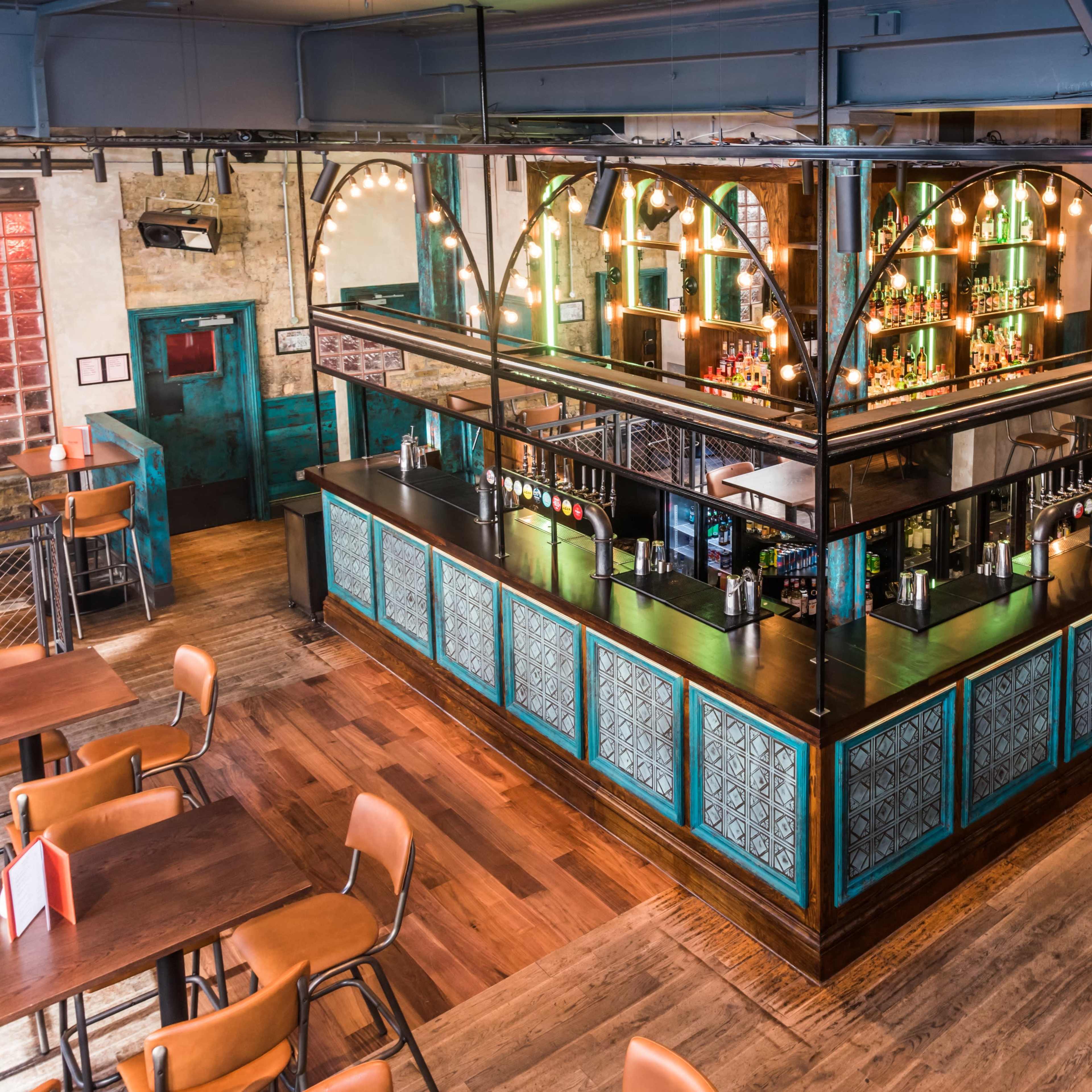 An elevated view of a modern bar area with a glass partition, illuminated shelves, and wooden flooring in a lively restaurant setting.
