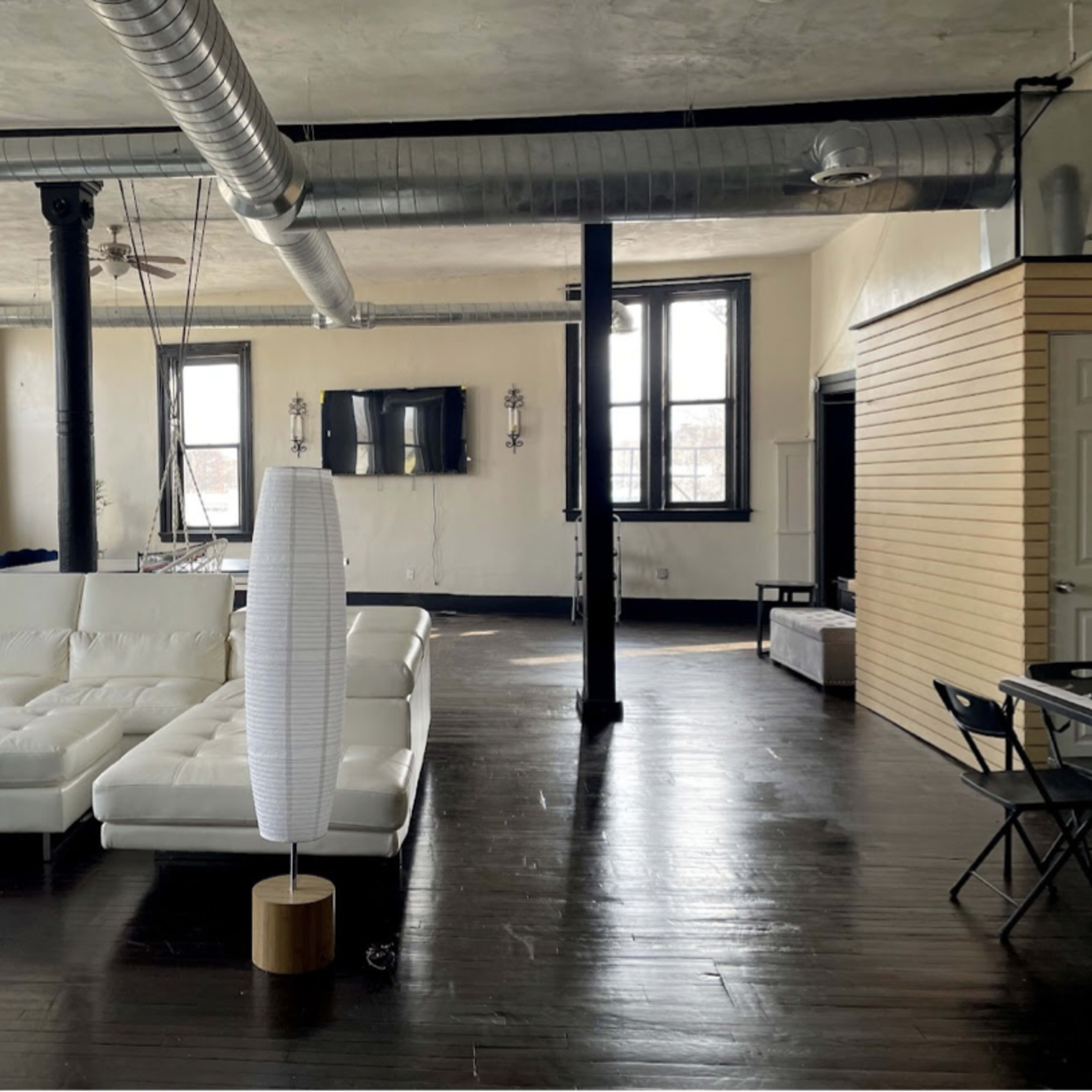 A spacious loft-style living area featuring a white sectional sofa, large windows, and exposed ductwork on the ceiling.