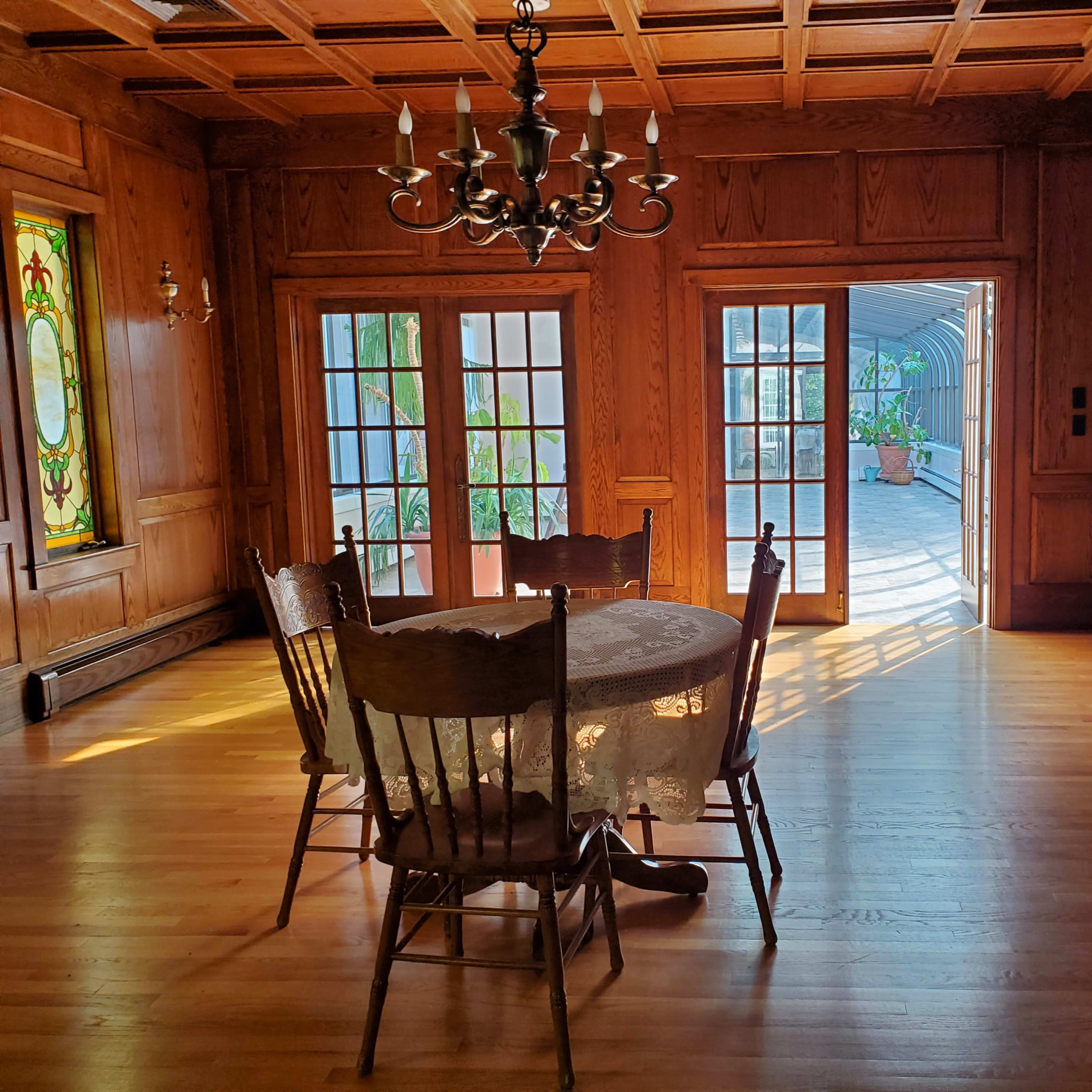 A wooden dining table surrounded by chairs is positioned in a room with paneled walls and large French doors leading to a patio.