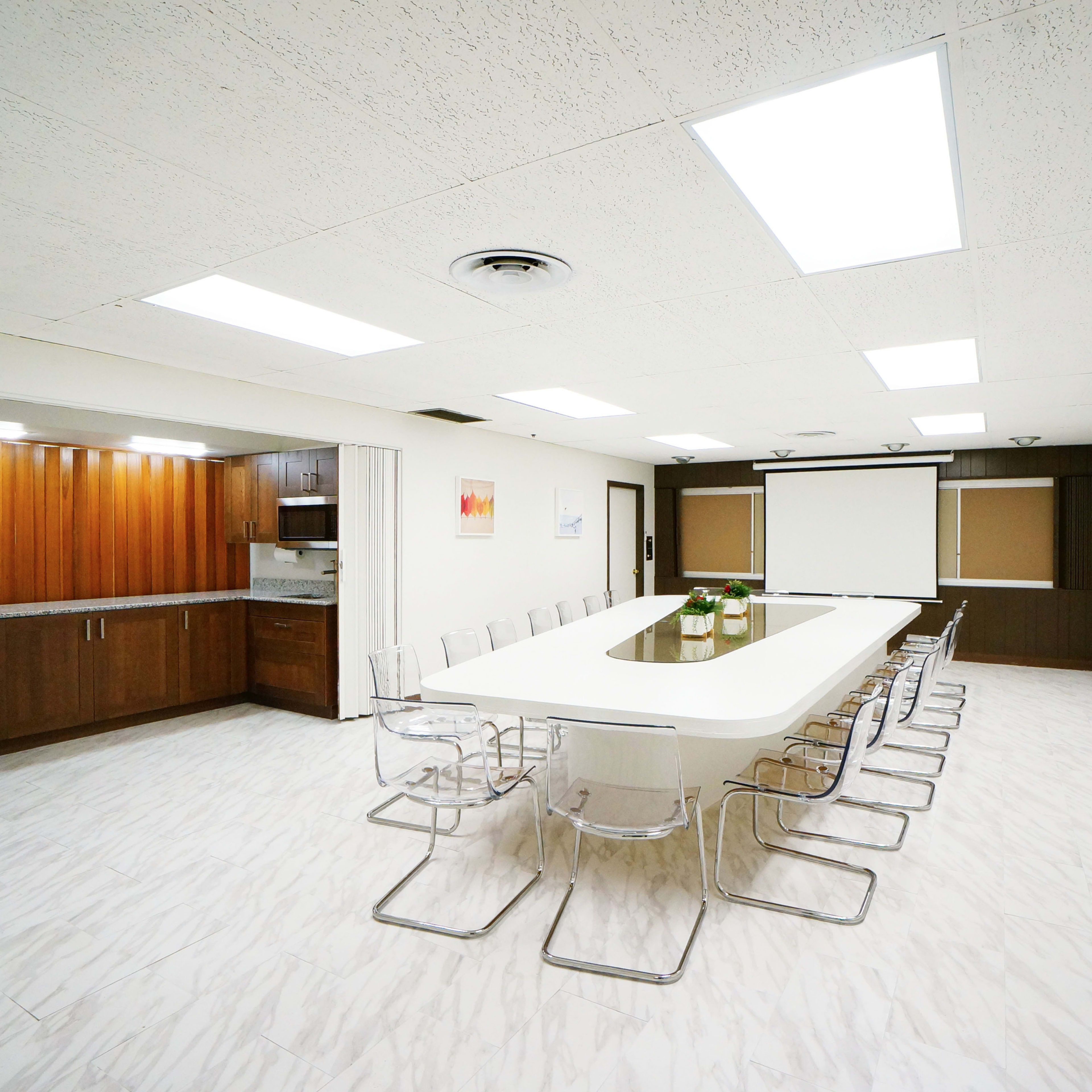 A modern conference room features a long table surrounded by clear chairs, with a kitchenette and a presentation screen in the background.