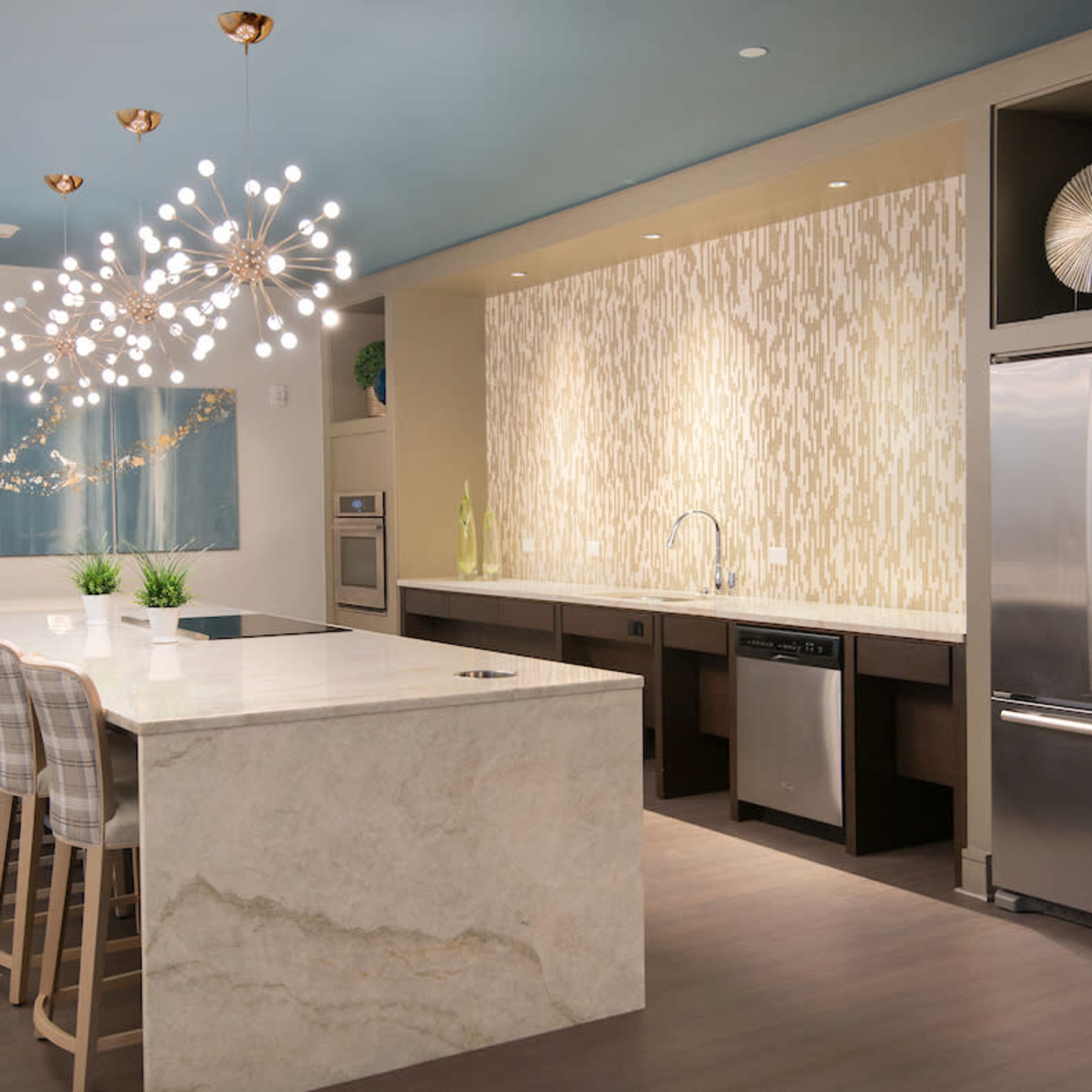 A modern kitchen features a large marble island, stainless steel appliances, and decorative lighting fixtures.