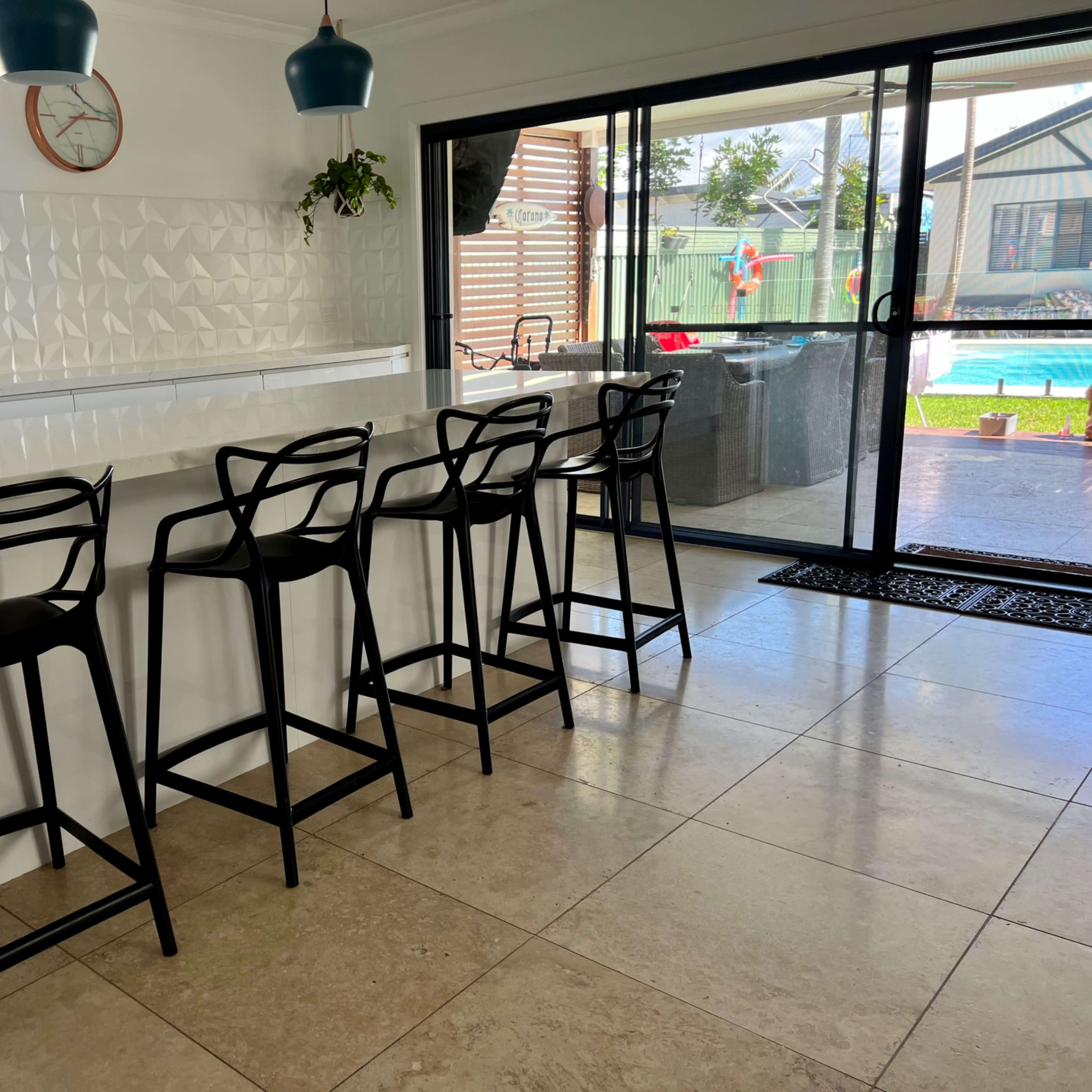 A modern kitchen features a sleek island with four black bar stools and a sliding glass door leading to an outdoor area with a swimming pool.