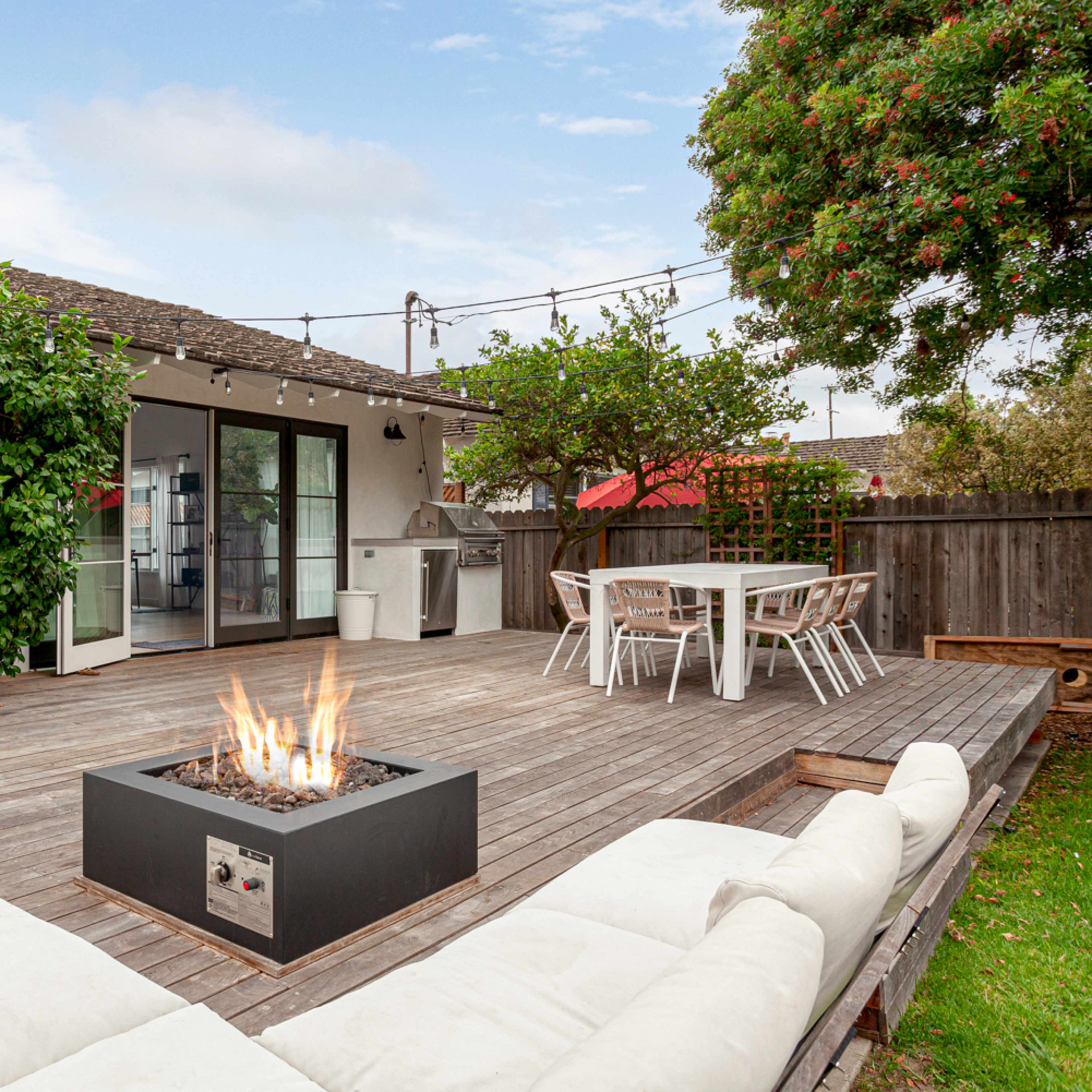 A wooden deck features a fire pit surrounded by cushioned seating, with a dining area and modern appliances in the background.