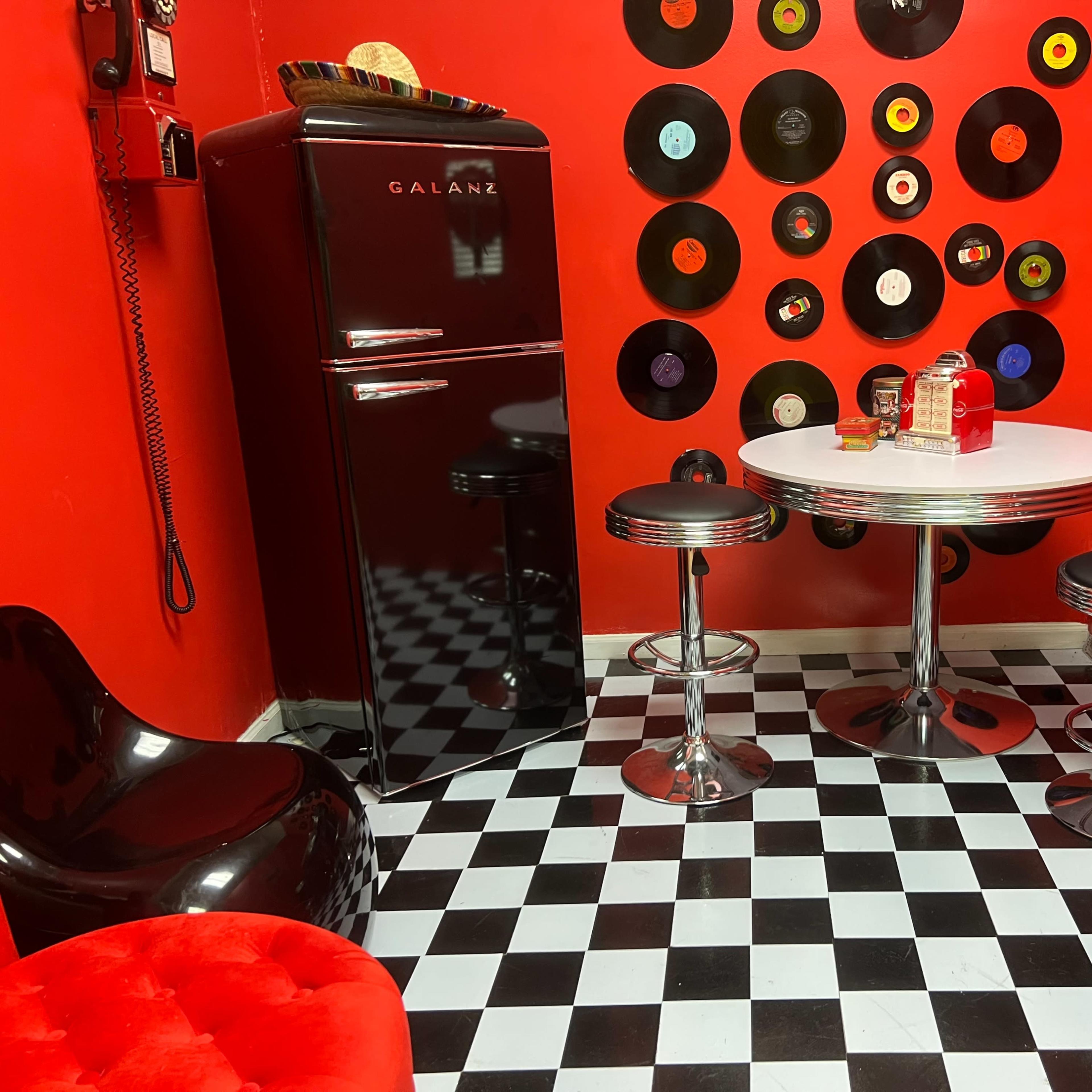 A retro-themed kitchen features a black refrigerator, a round white table with chrome stools, and a wall adorned with vinyl records against a red backdrop.