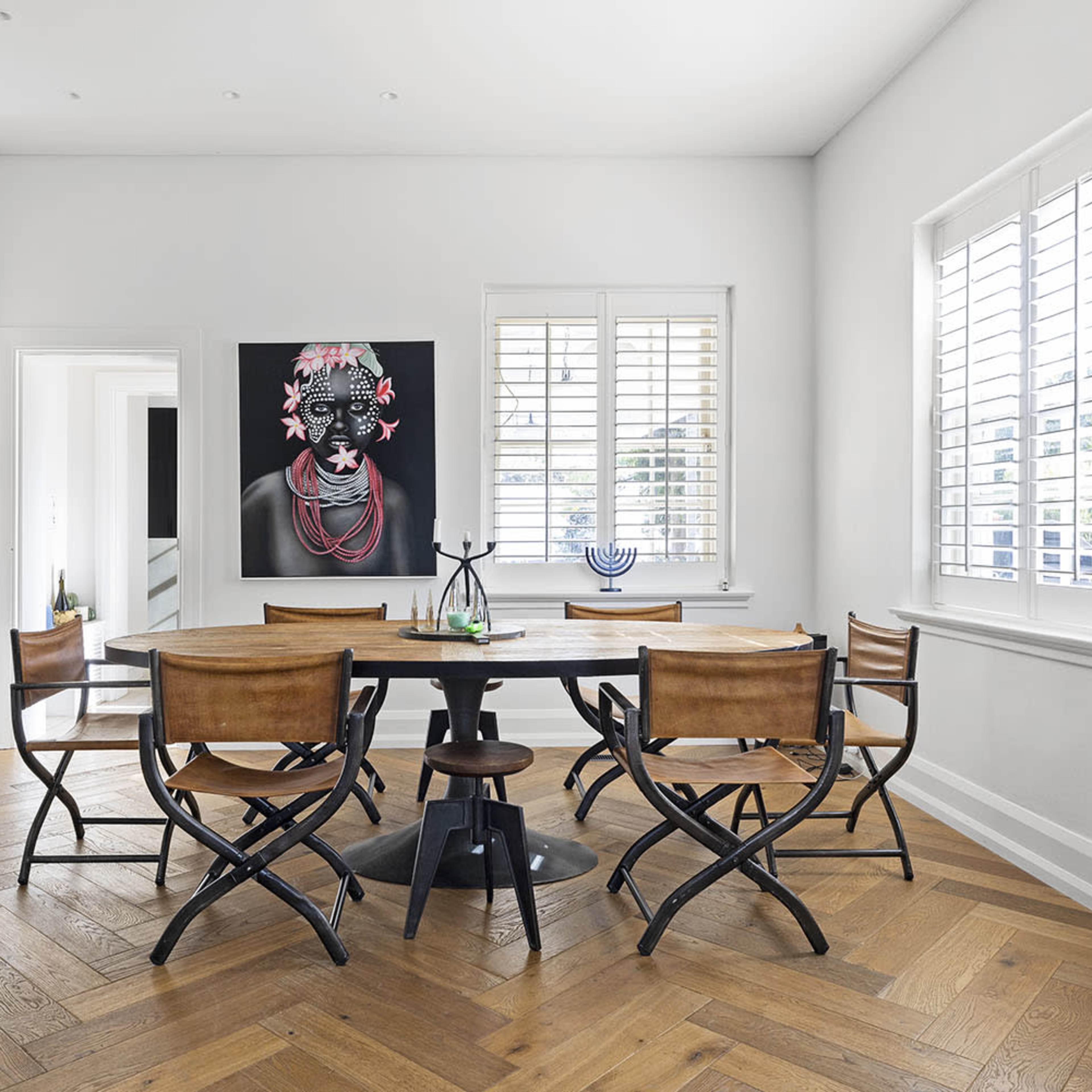 A modern dining area featuring a round table surrounded by several chairs, with two large framed artworks on the wall and a shelf displaying various decor items.