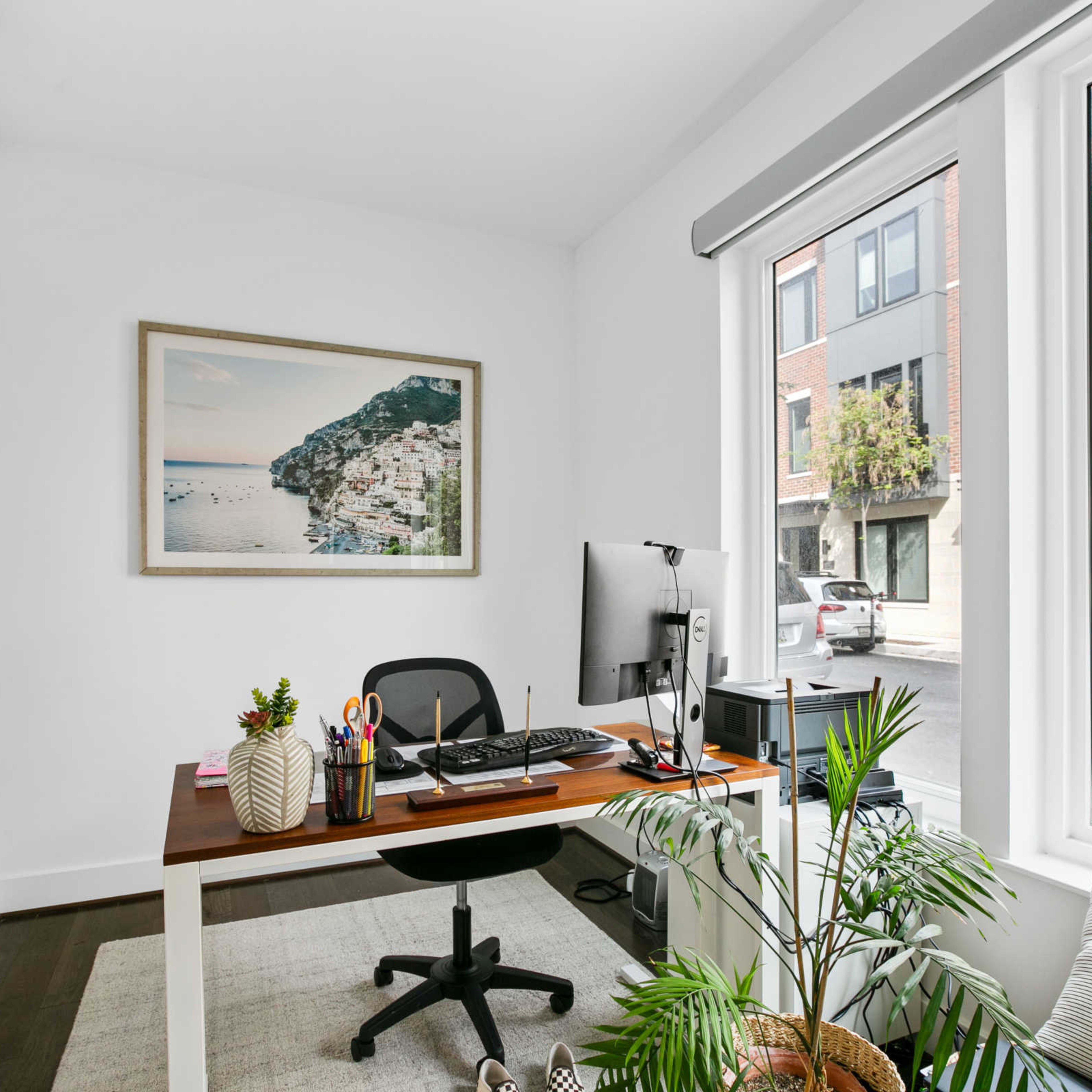 A home office features a desk with a computer and office supplies, alongside a window with a view of a street and greenery.