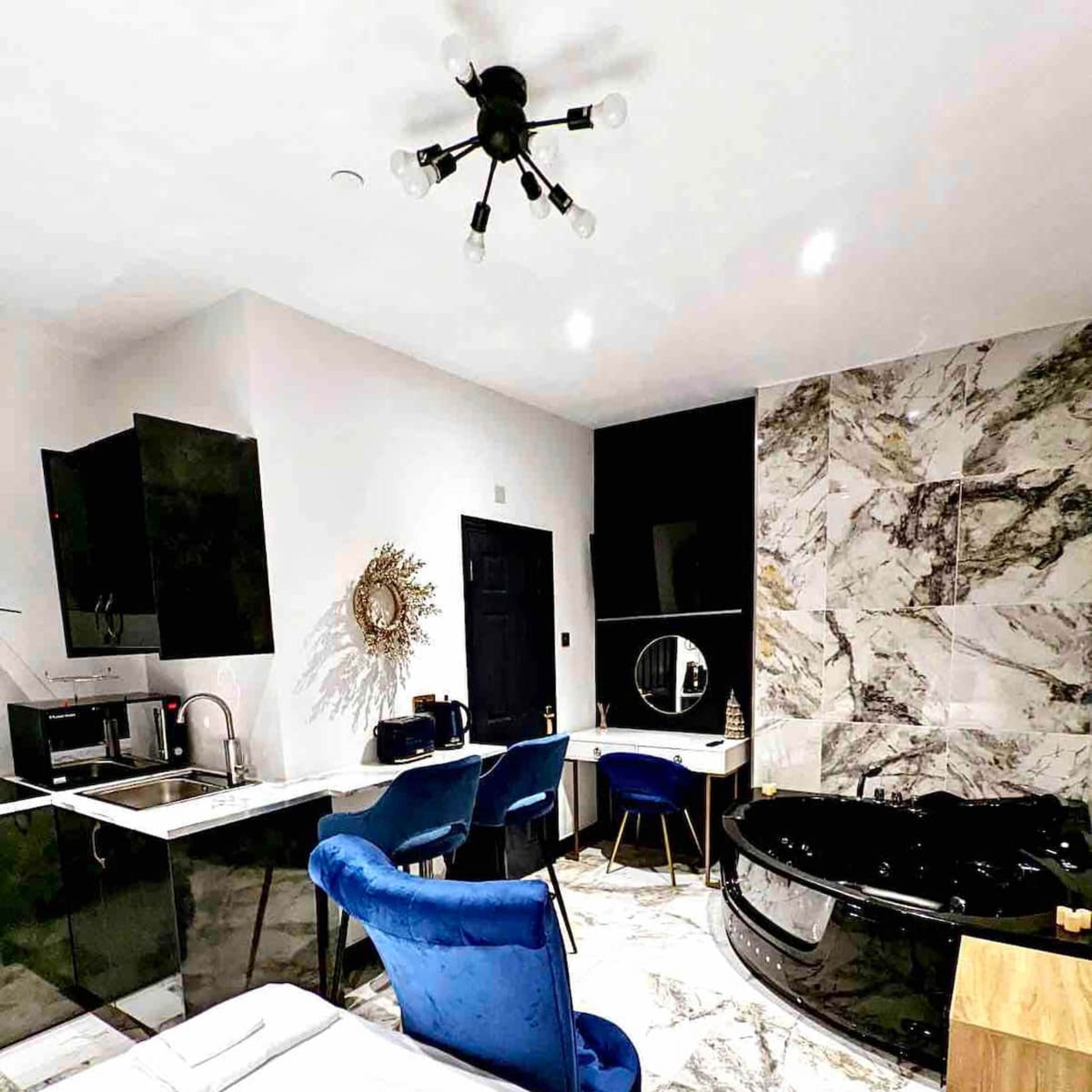 A modern hotel room featuring a bed, a kitchen area with black cabinetry, a small dining table with blue chairs, and a black whirlpool tub.