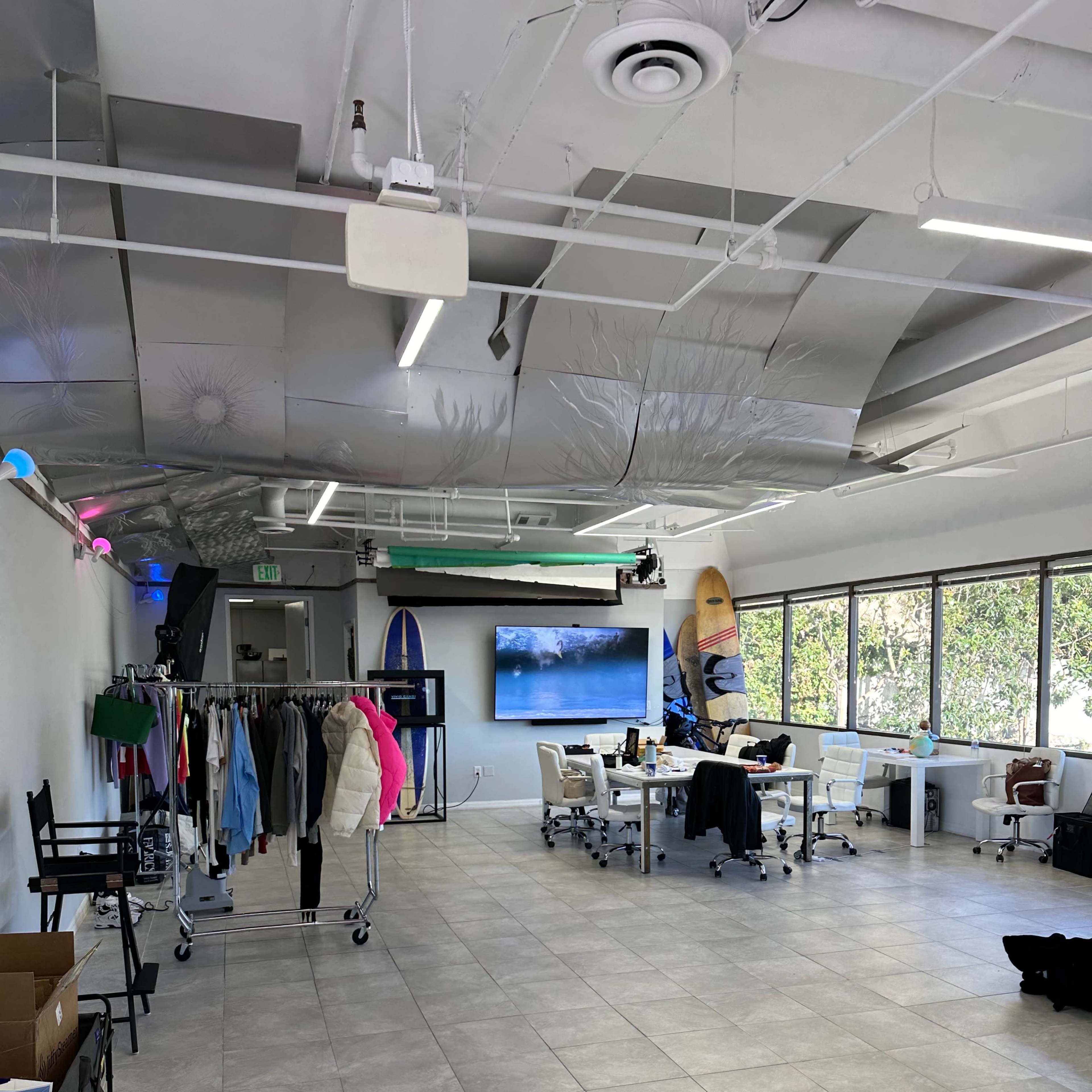 A spacious office area features clothing racks, workstations, and surfboards displayed against a wall of large windows.