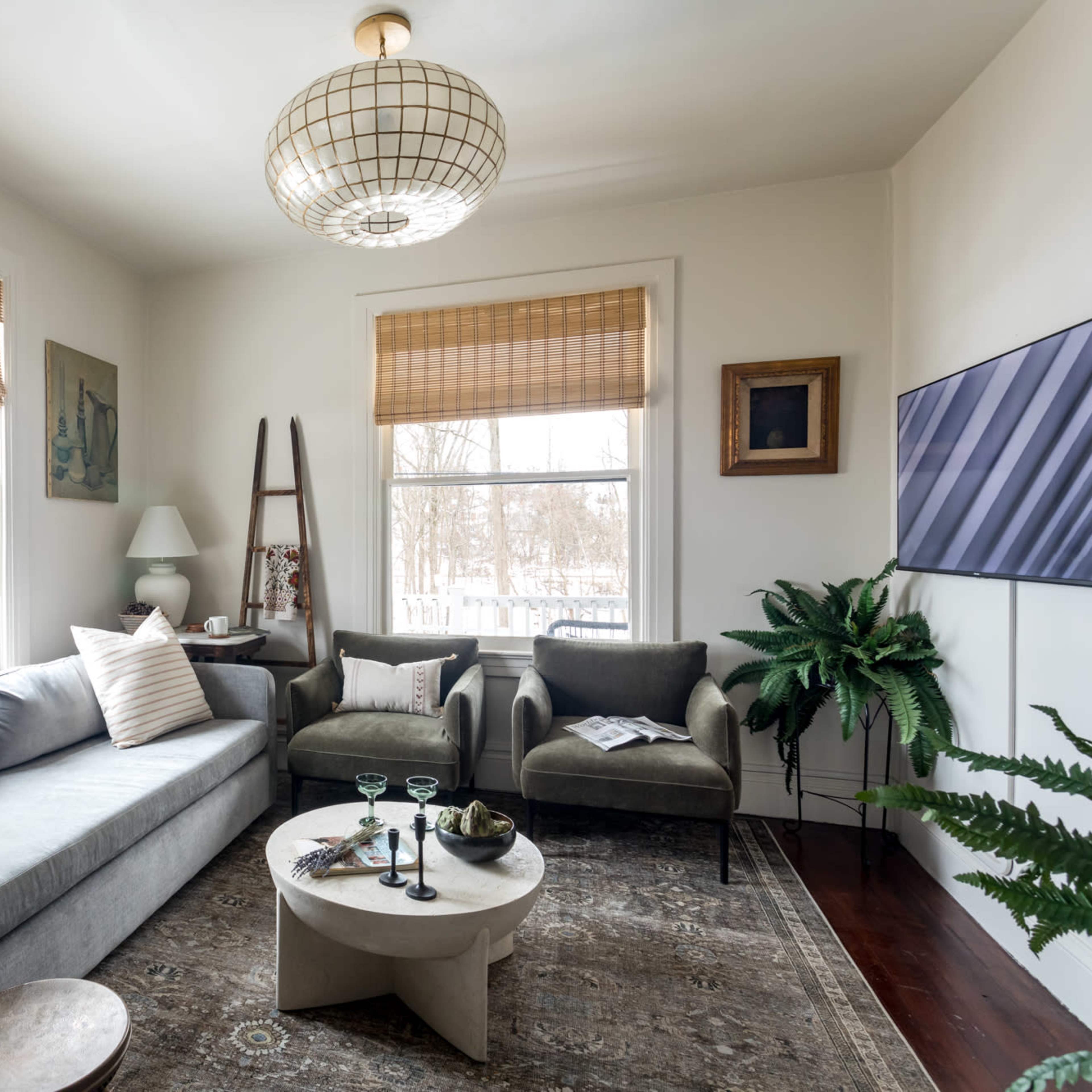 A cozy living room features a gray sofa, two green chairs, a round coffee table, and large windows with bamboo shades, surrounded by plants.