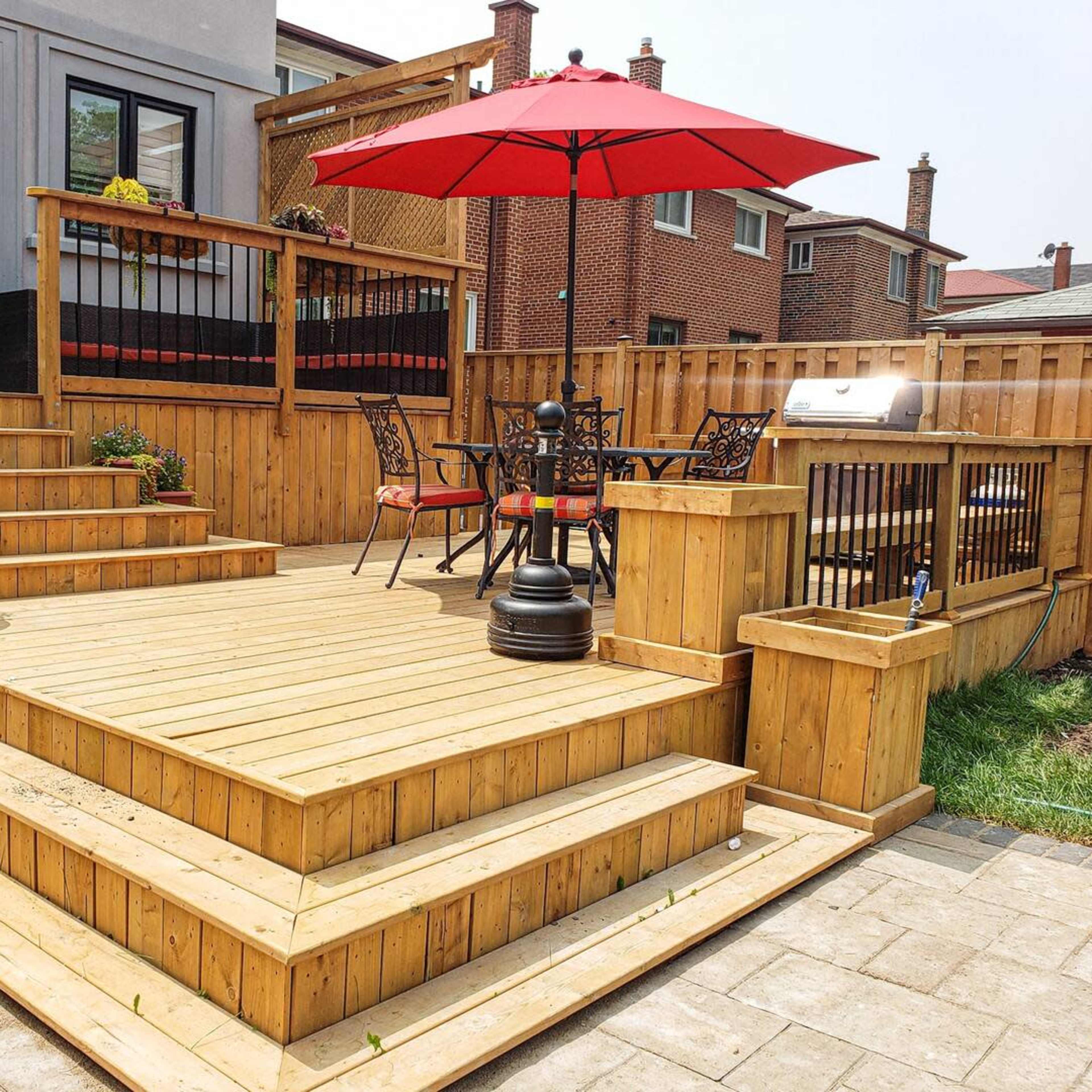 A wooden deck with steps, an outdoor table and chairs under a red umbrella, and a grill surrounded by a fenced backyard.