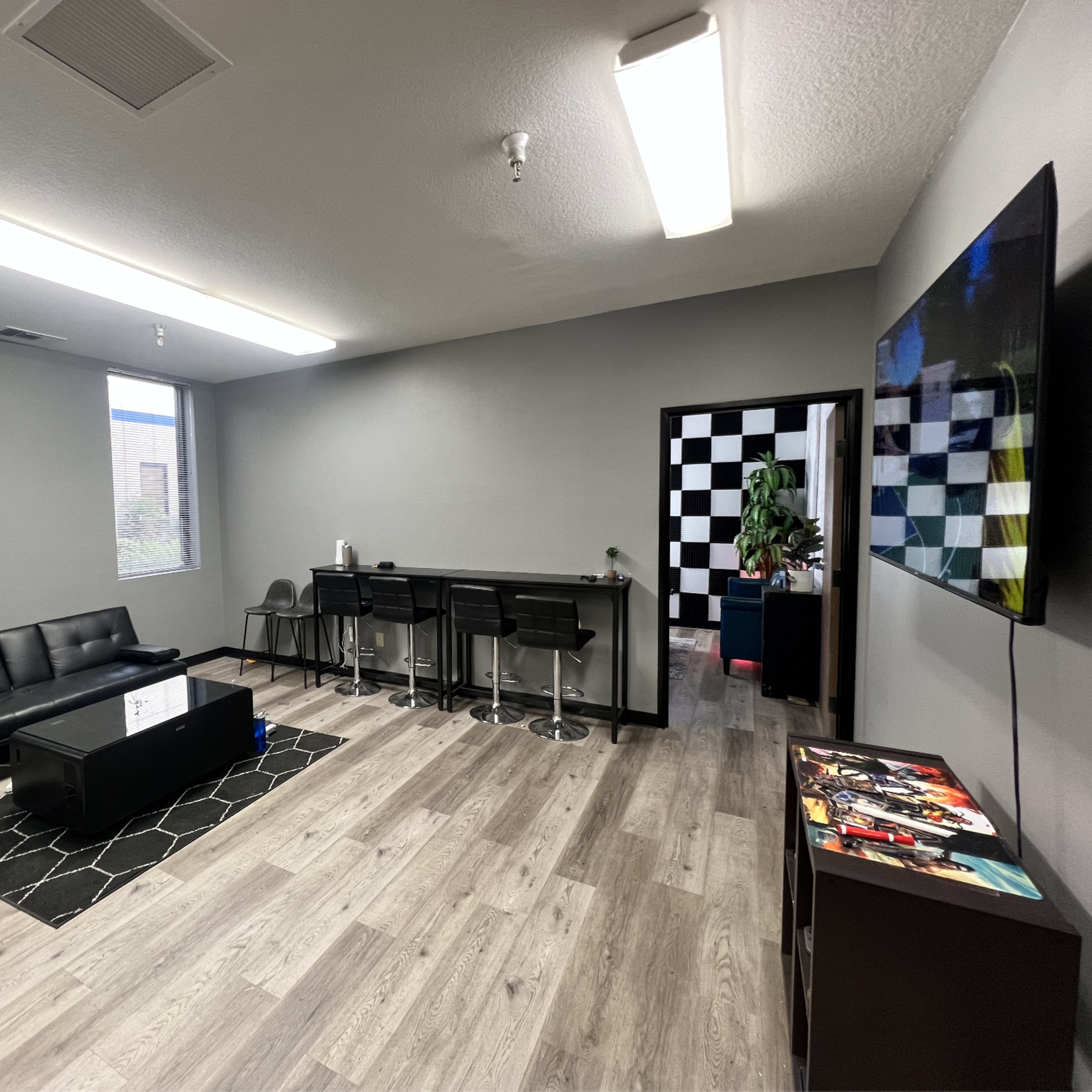 A modern lounge area with a black sofa, barstools along a counter, a large TV on the wall, and a patterned floor.
