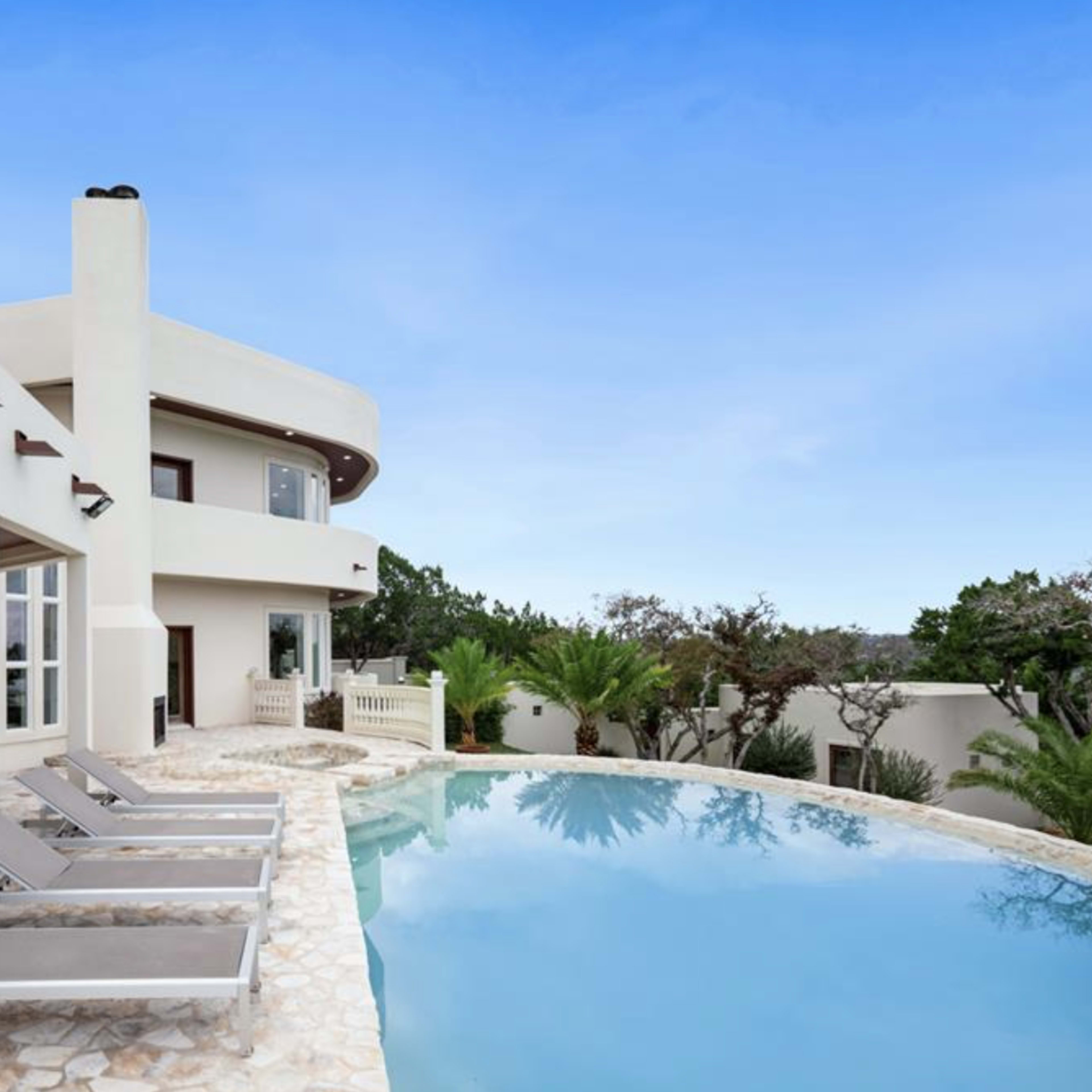 A modern villa with a curved design features a stone patio, lounge chairs, and a pool surrounded by lush greenery.