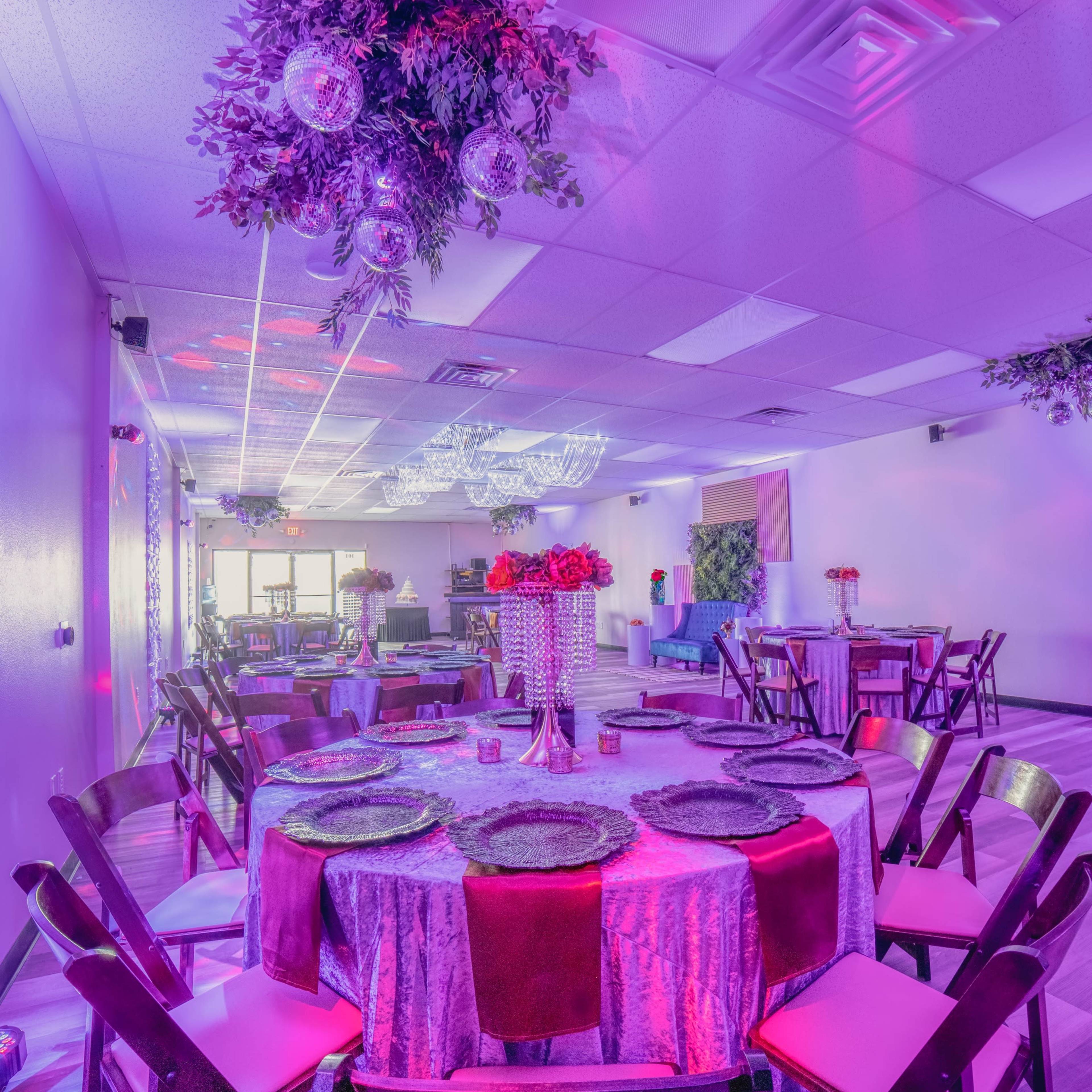A brightly lit event space features round tables set with colorful tableware and floral arrangements under decorative lighting.