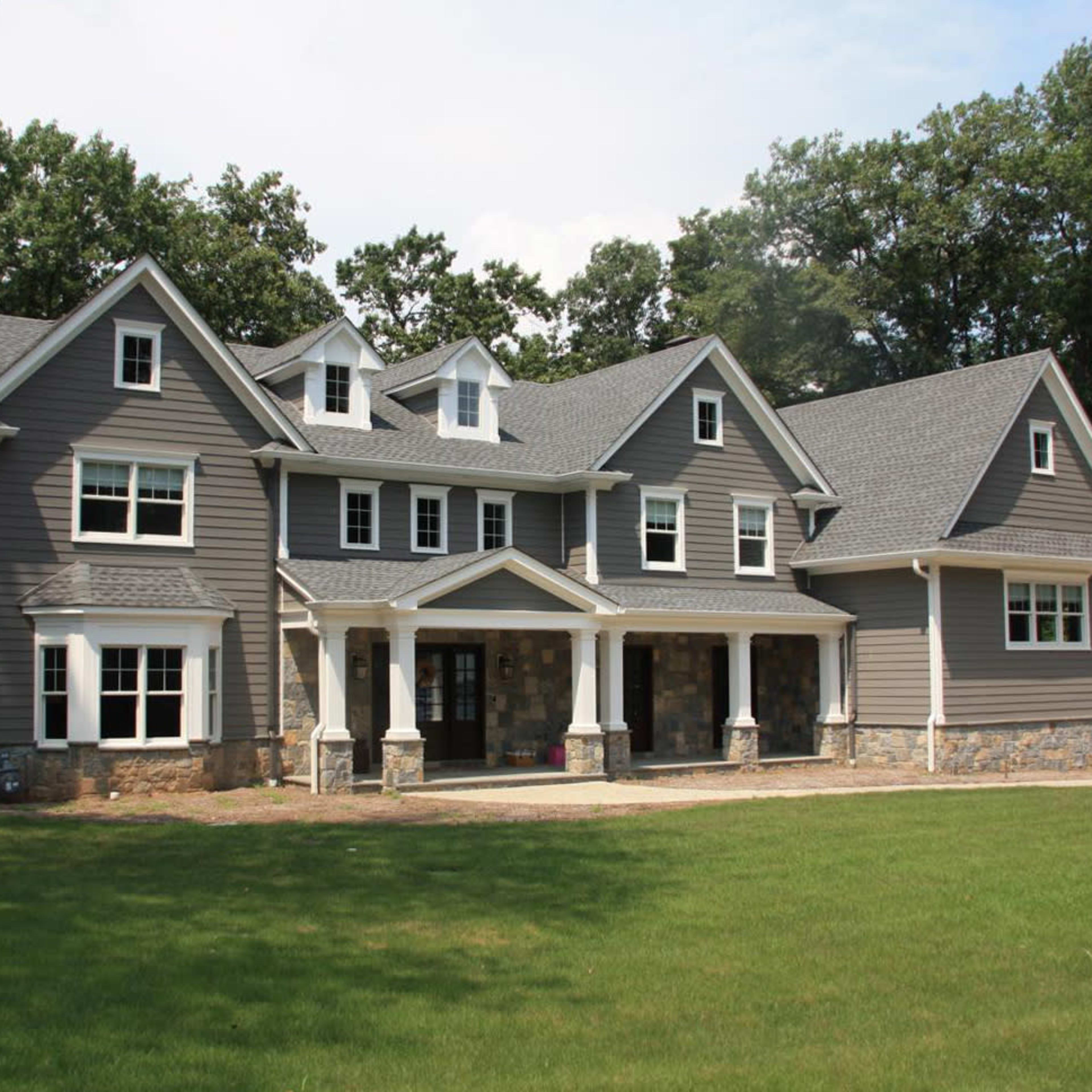 A large gray multi-story house with a stone foundation and multiple gabled roofs is set on a grassy lawn surrounded by trees.