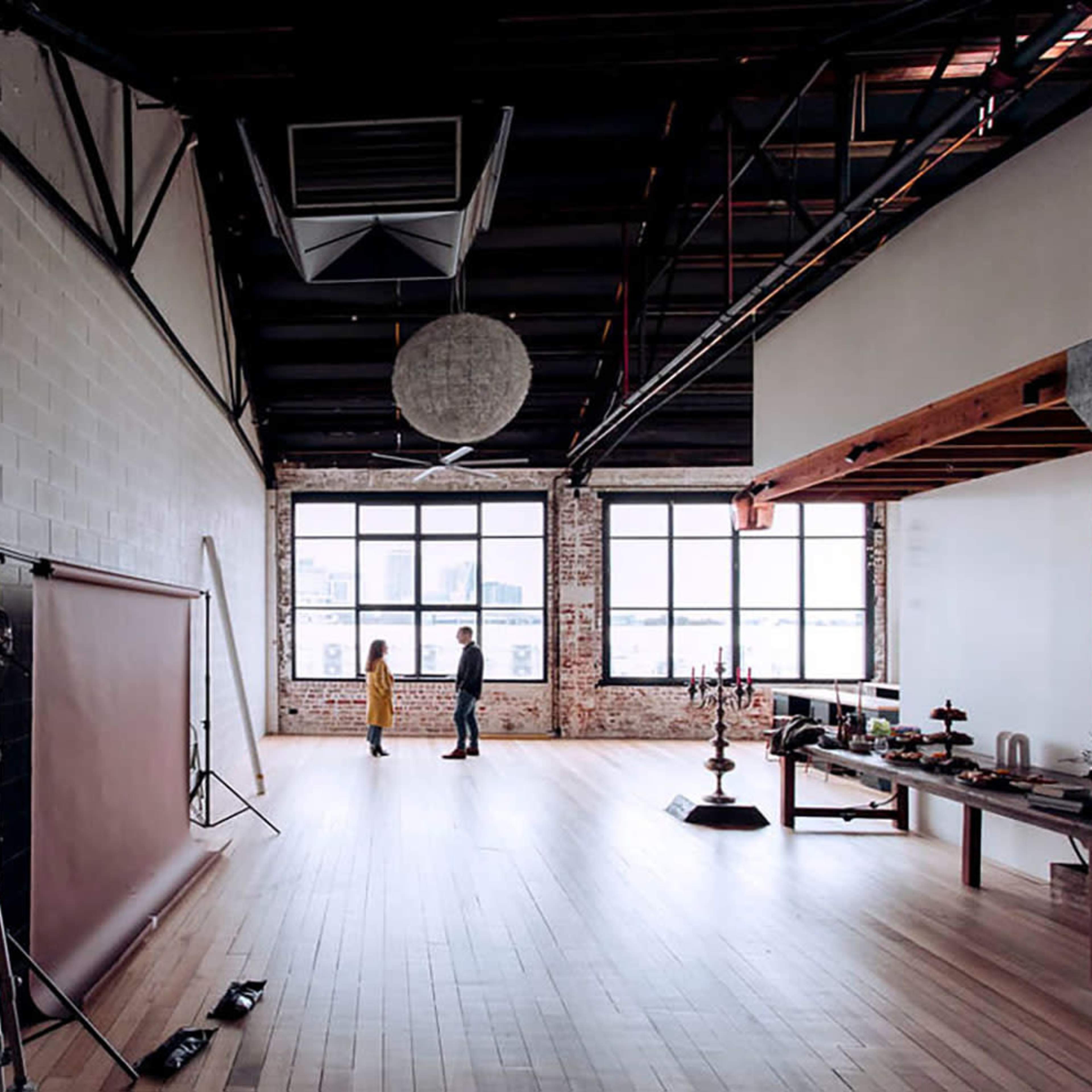 A spacious industrial studio with large windows, a smooth wooden floor, and a few scattered props, including a backdrop, lighting equipment, and a table with various items.
