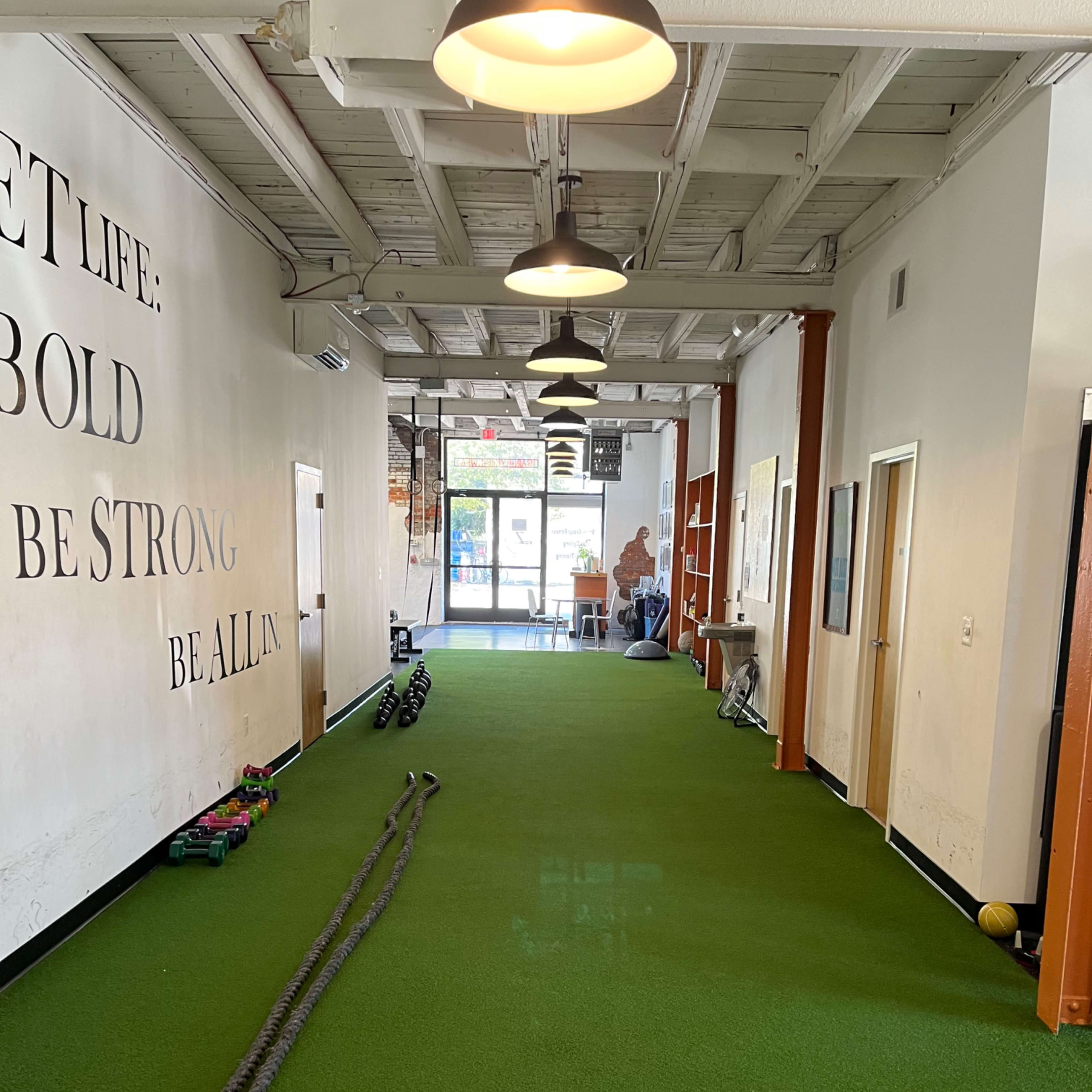 A long hallway with green turf flooring features workout equipment such as ropes and weights along the sides, leading to a glass entrance at the far end.