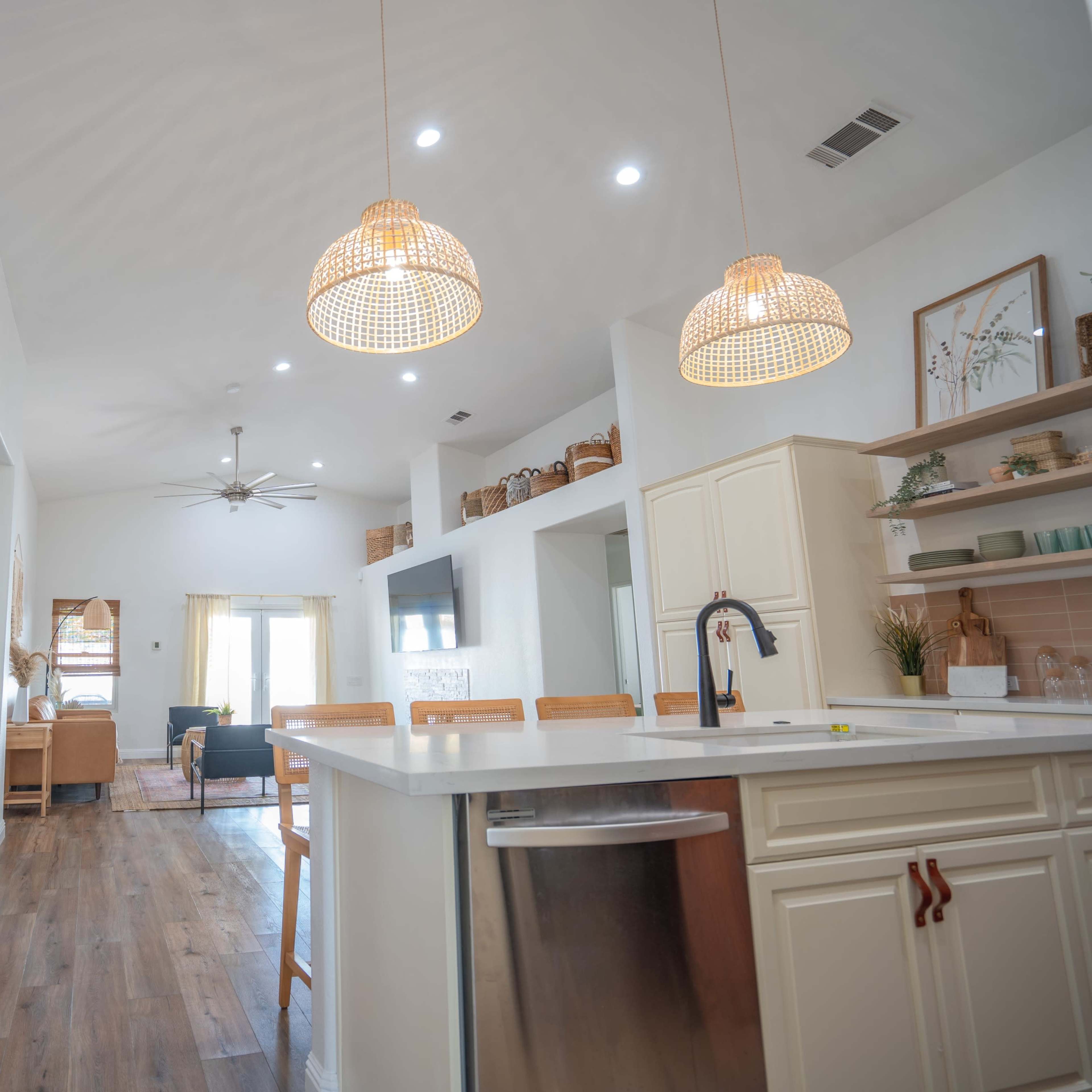 A modern kitchen with a white island, pendant lights, and a large mural featuring stylized plant leaves, leading into a living space with natural light.