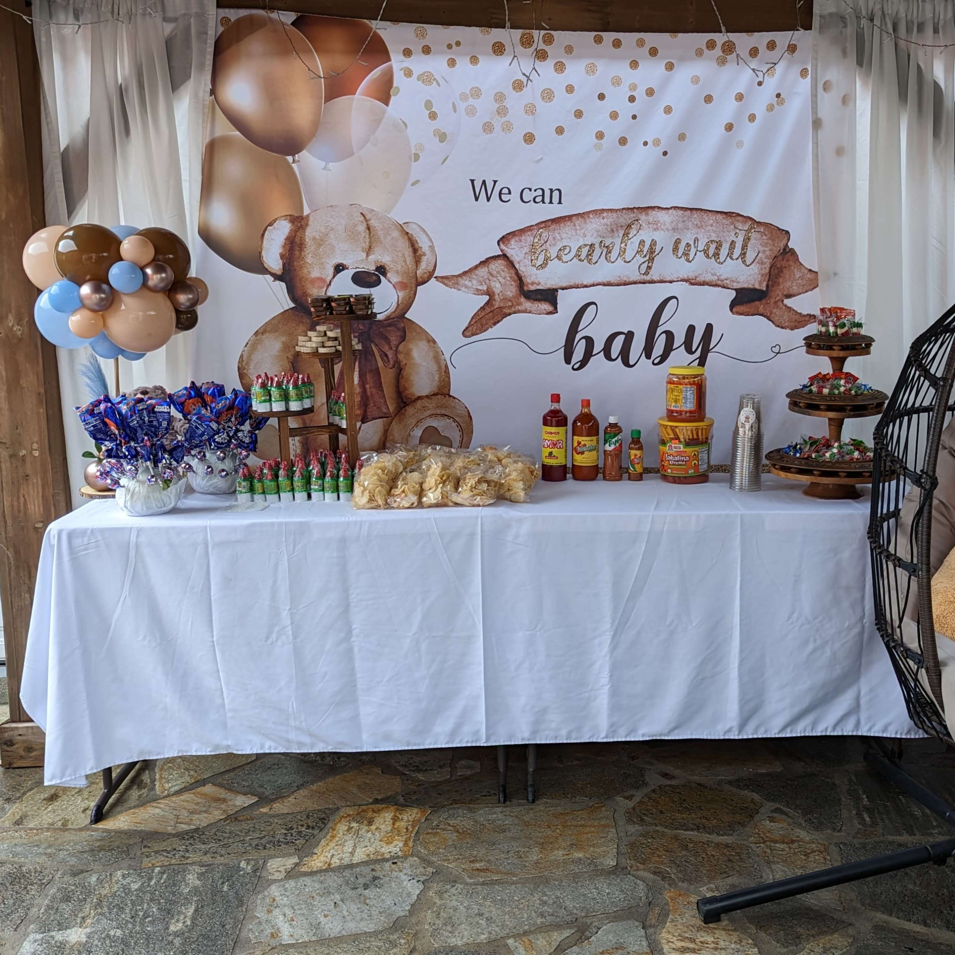 A decorated table with snacks and beverages, featuring a teddy bear theme, under a canopy with a large banner that says "We can bear-ly wait baby."