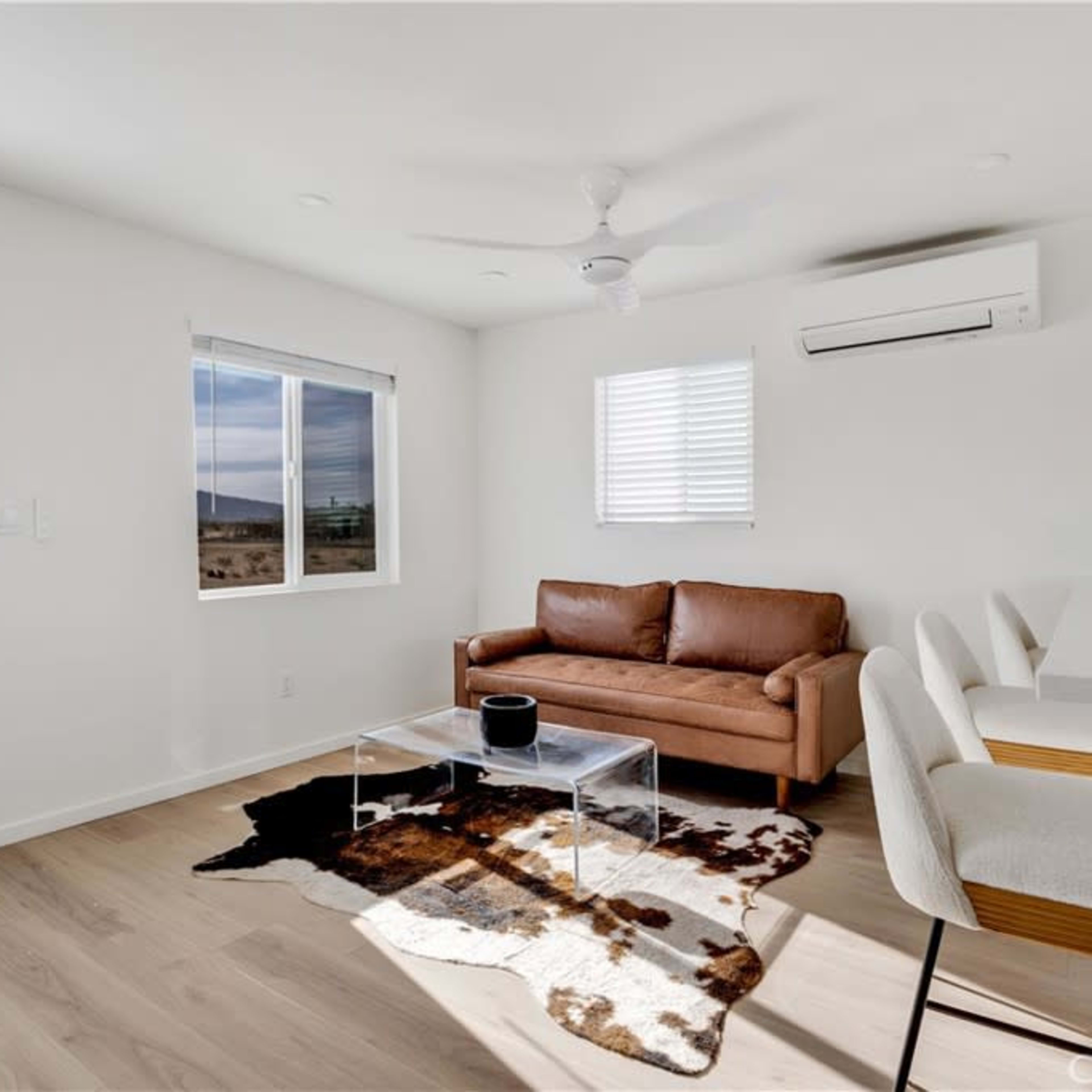 A bright, modern living room with a brown leather couch, a clear coffee table, and a small dining area featuring white stools.