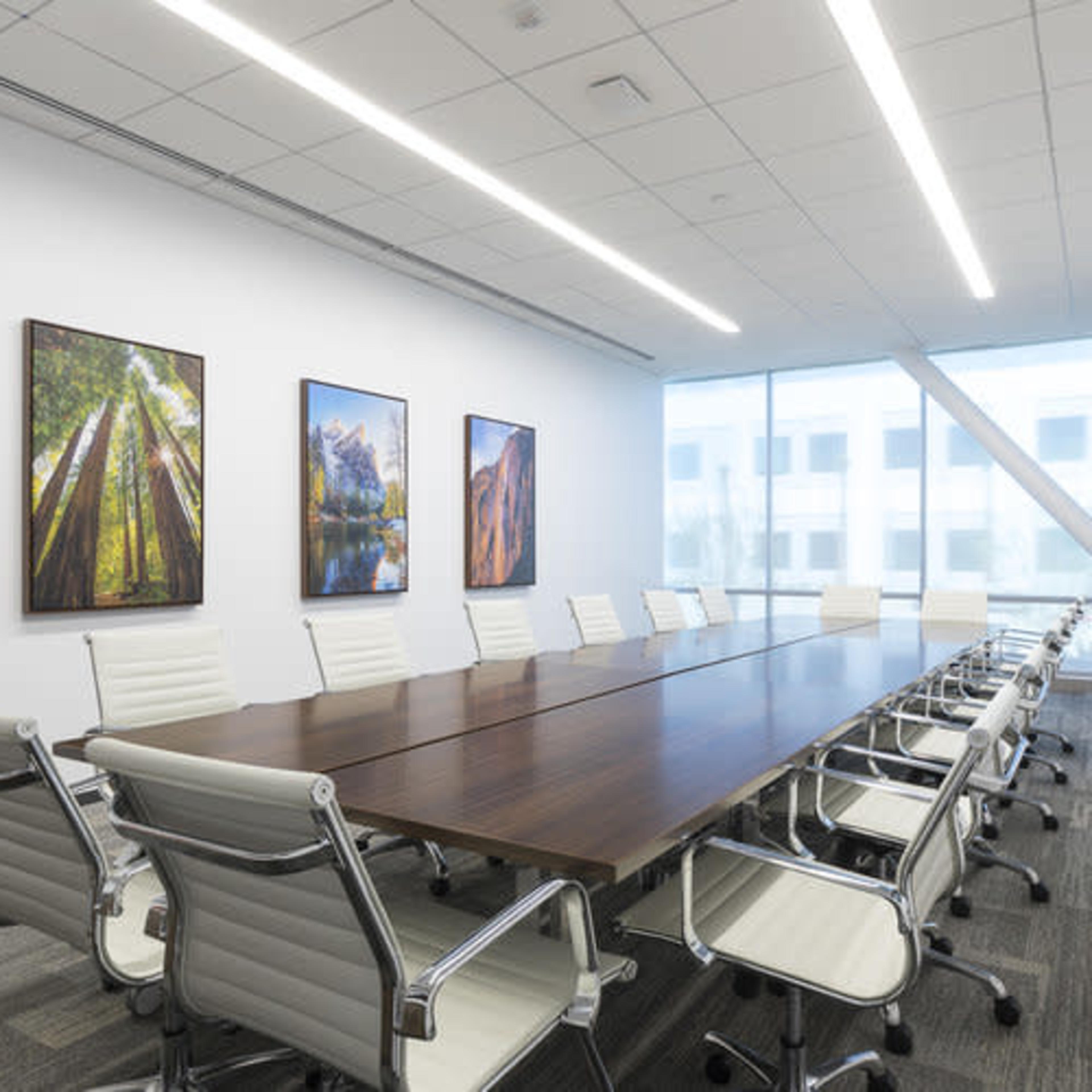 A modern conference room featuring a long wooden table surrounded by white chairs and large windows displaying artwork on the walls.