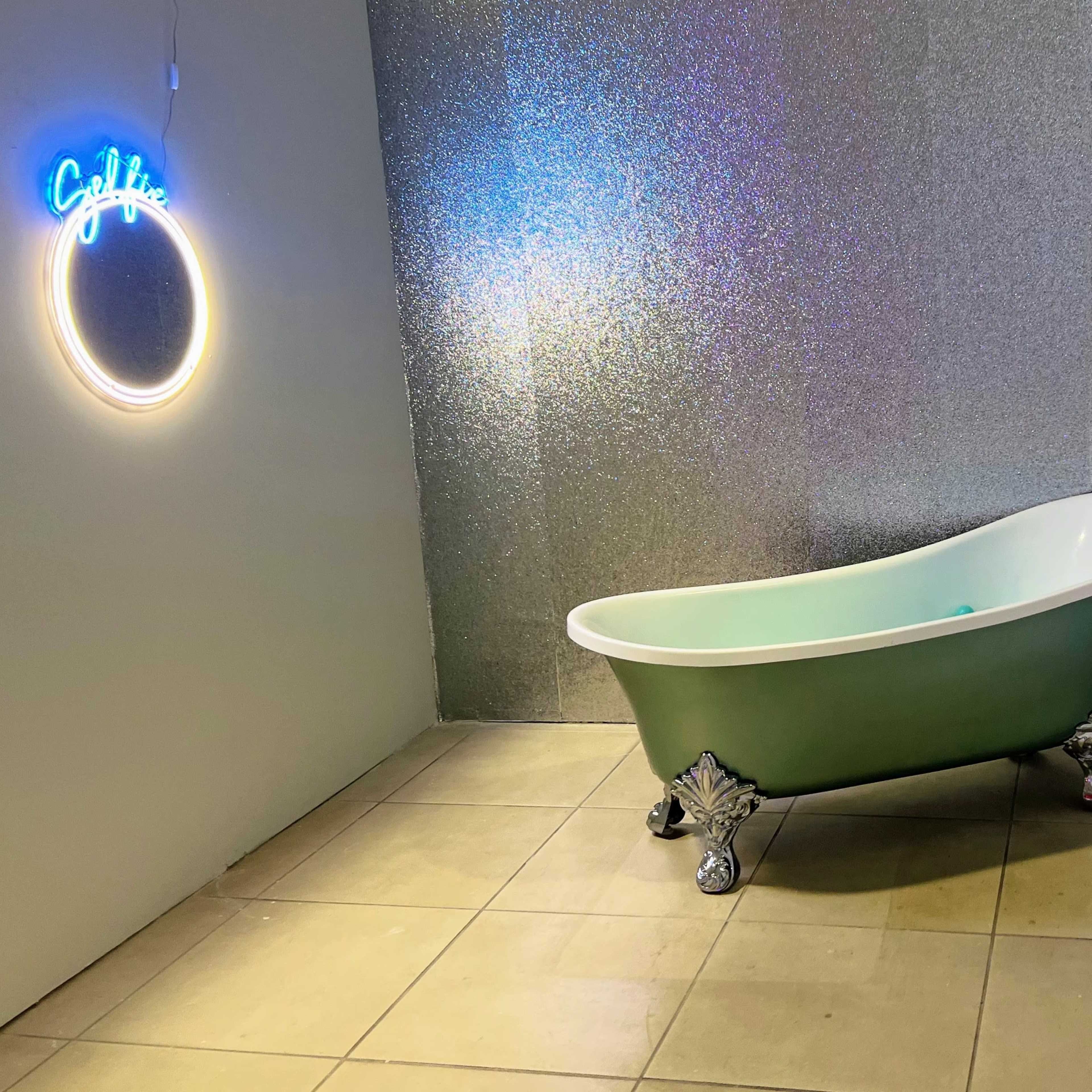A vintage-style green bathtub with ornate silver feet is placed against a shimmering silver wall, accompanied by a neon sign that reads "Coffee."