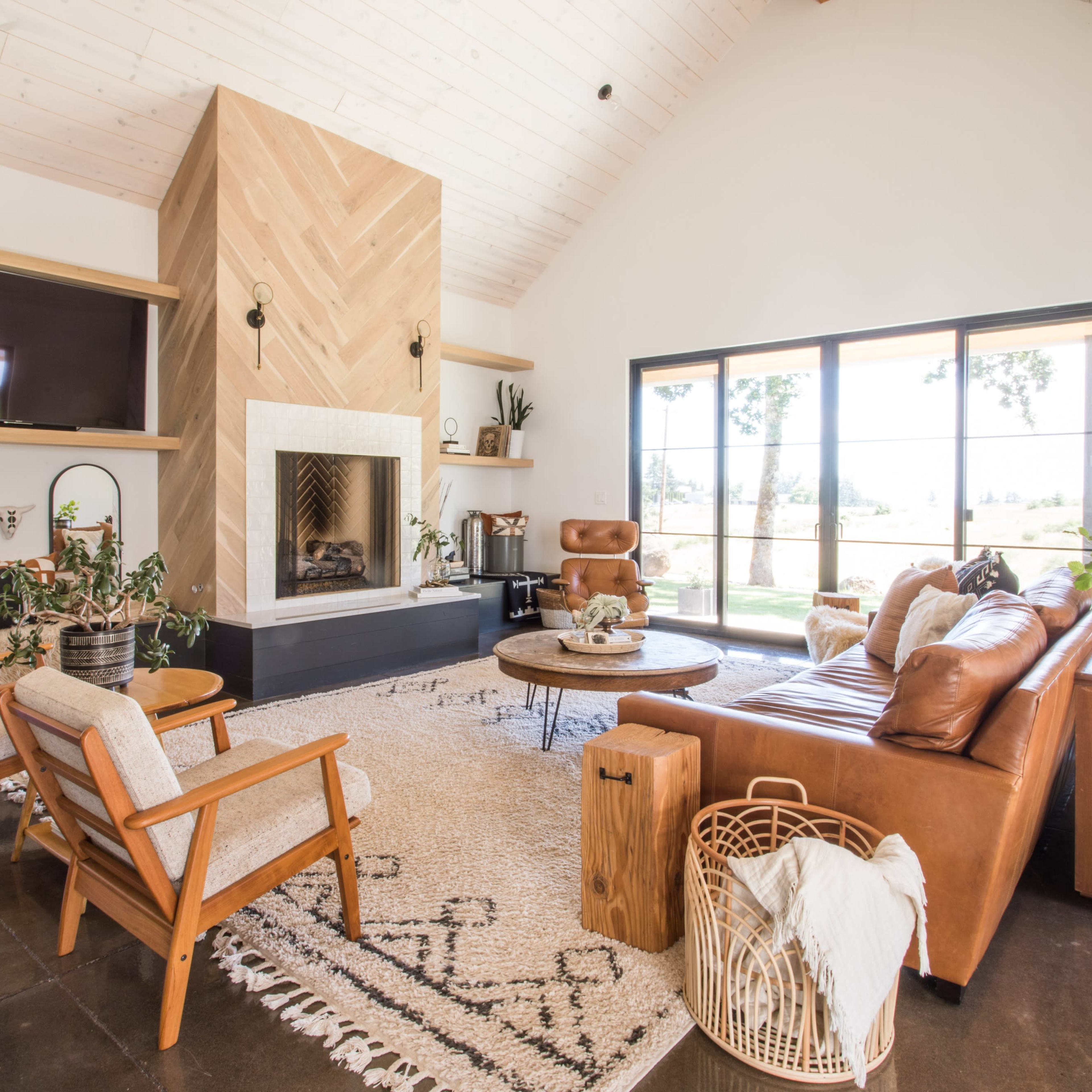 A modern living room with a large fireplace, a leather sofa, wooden chairs, and glass doors that lead to an outdoor view.