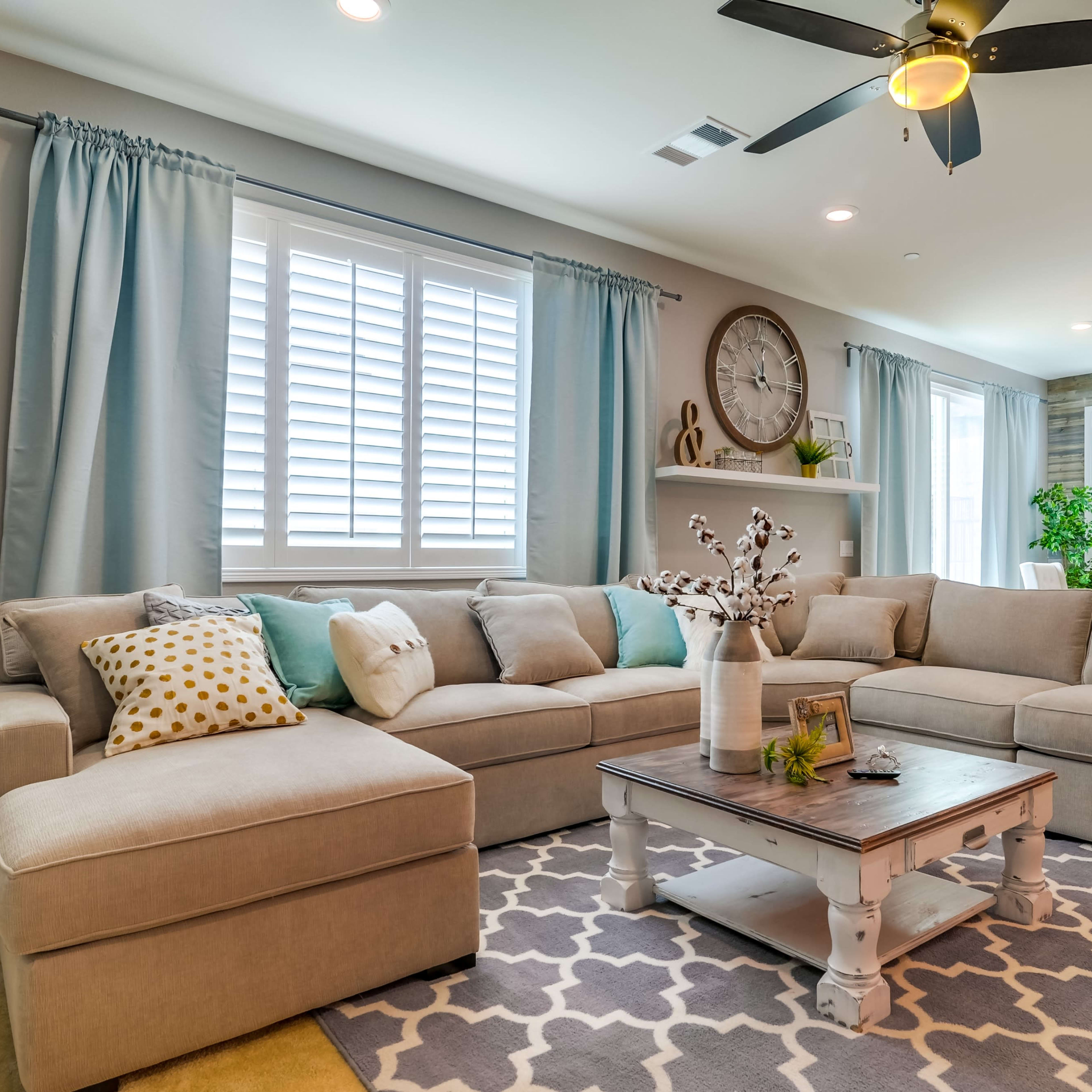A spacious living room features a beige sectional sofa, a white coffee table, and large windows with light blue curtains.