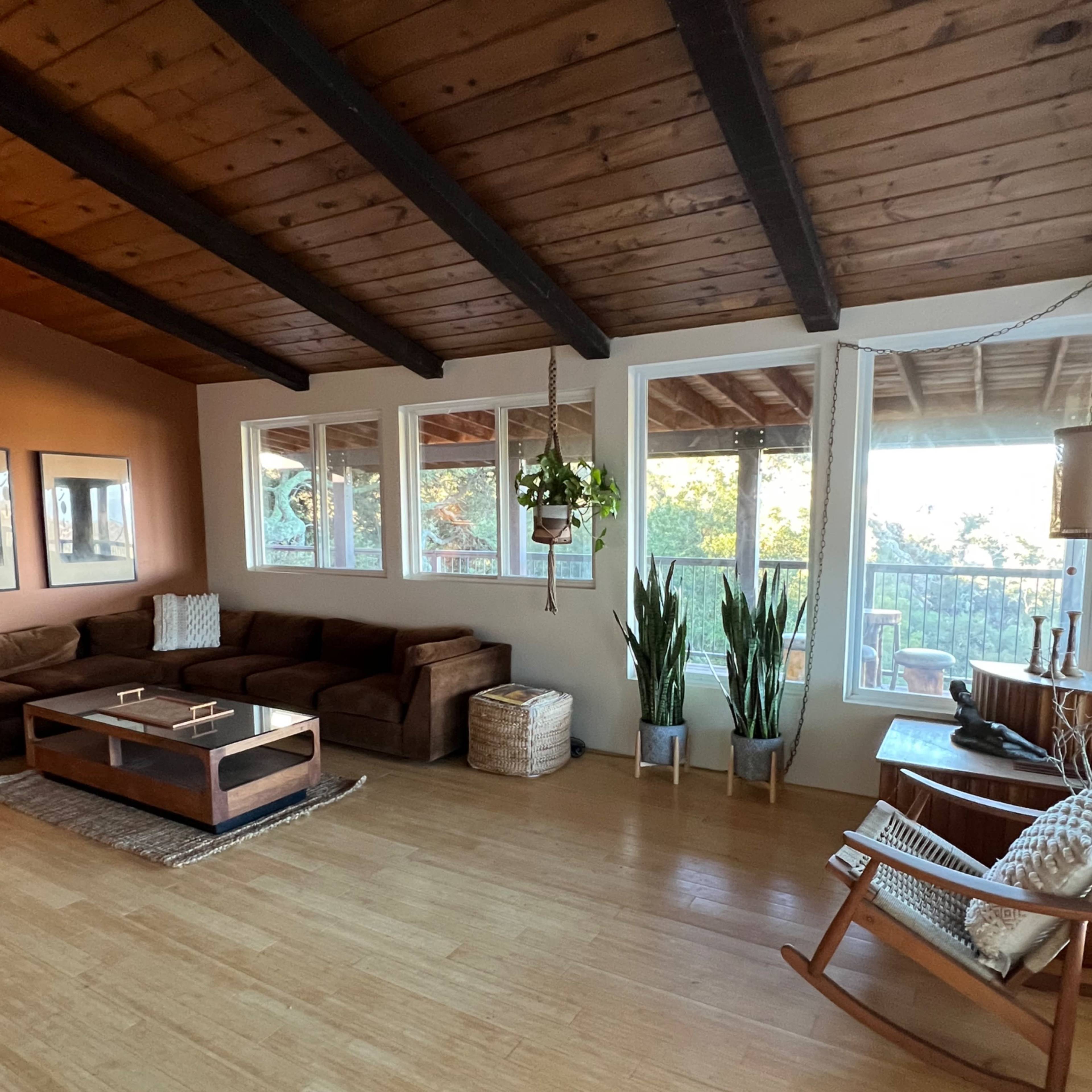 The living room features a brown sectional sofa, a coffee table, large windows with views outside, and indoor plants beside a rocking chair.
