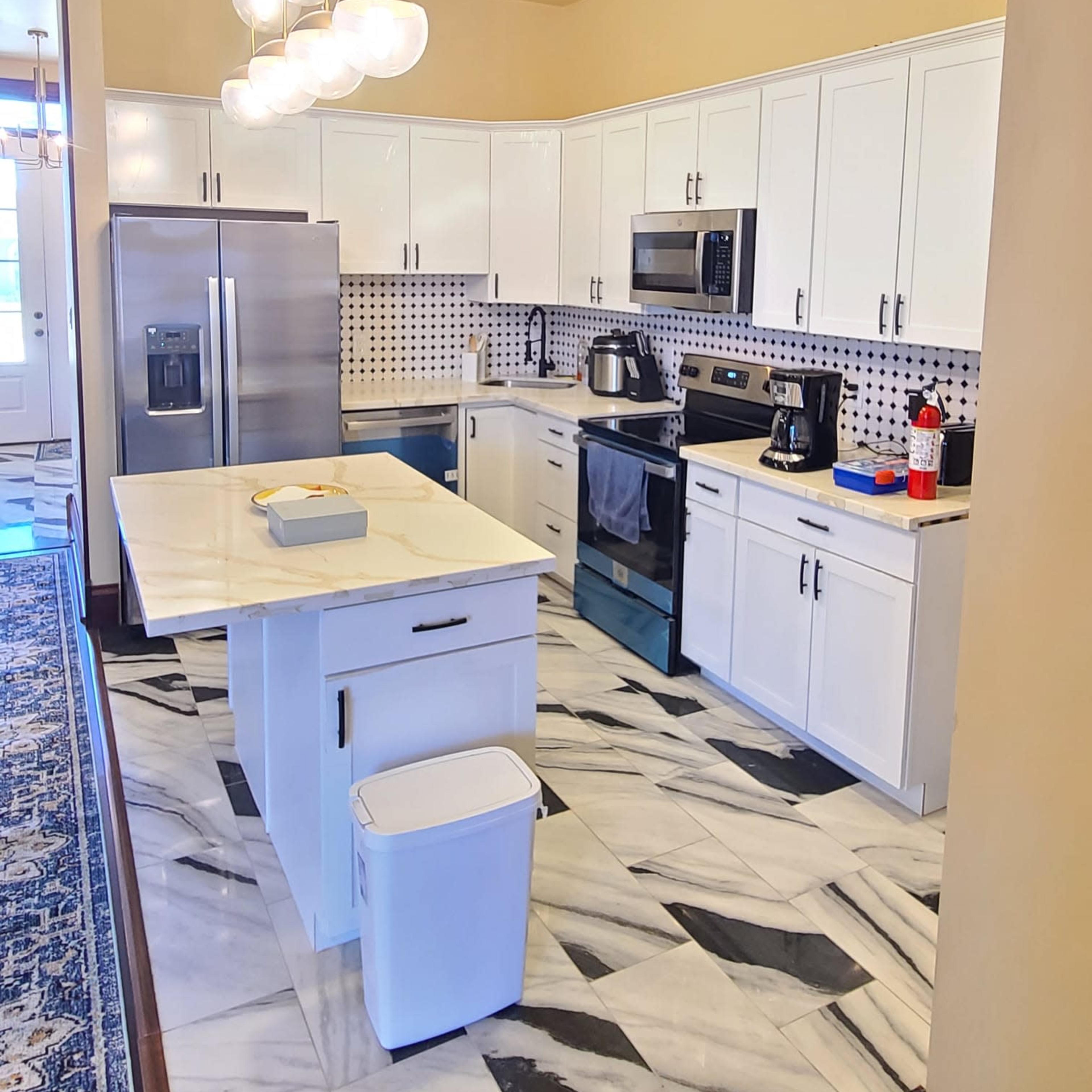 A modern kitchen features white cabinets, stainless steel appliances, and a marble-patterned floor, with a central island and pendant lighting above.