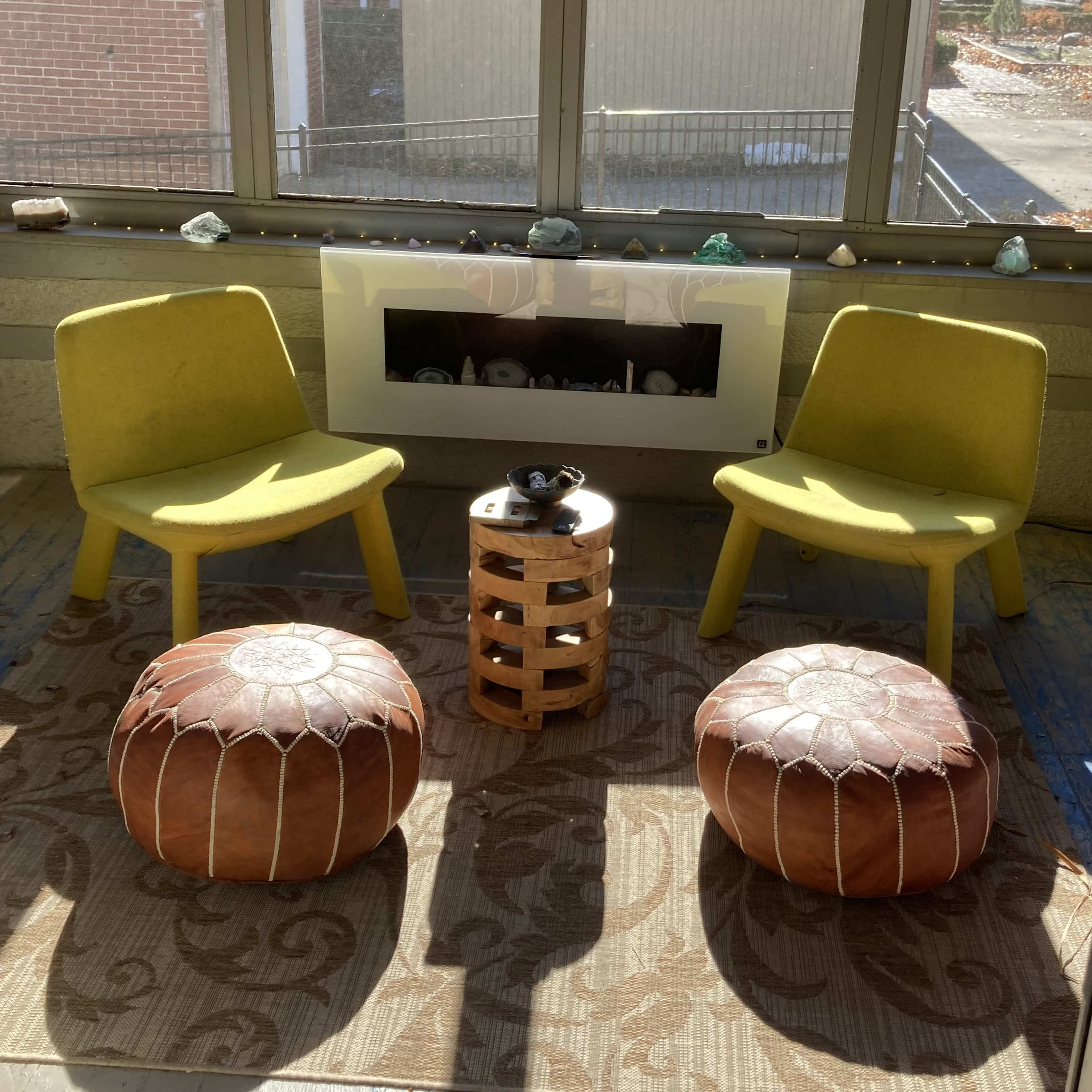 A cozy sunlit seating area with two yellow chairs, a wooden table, and two brown poufs on a patterned rug.