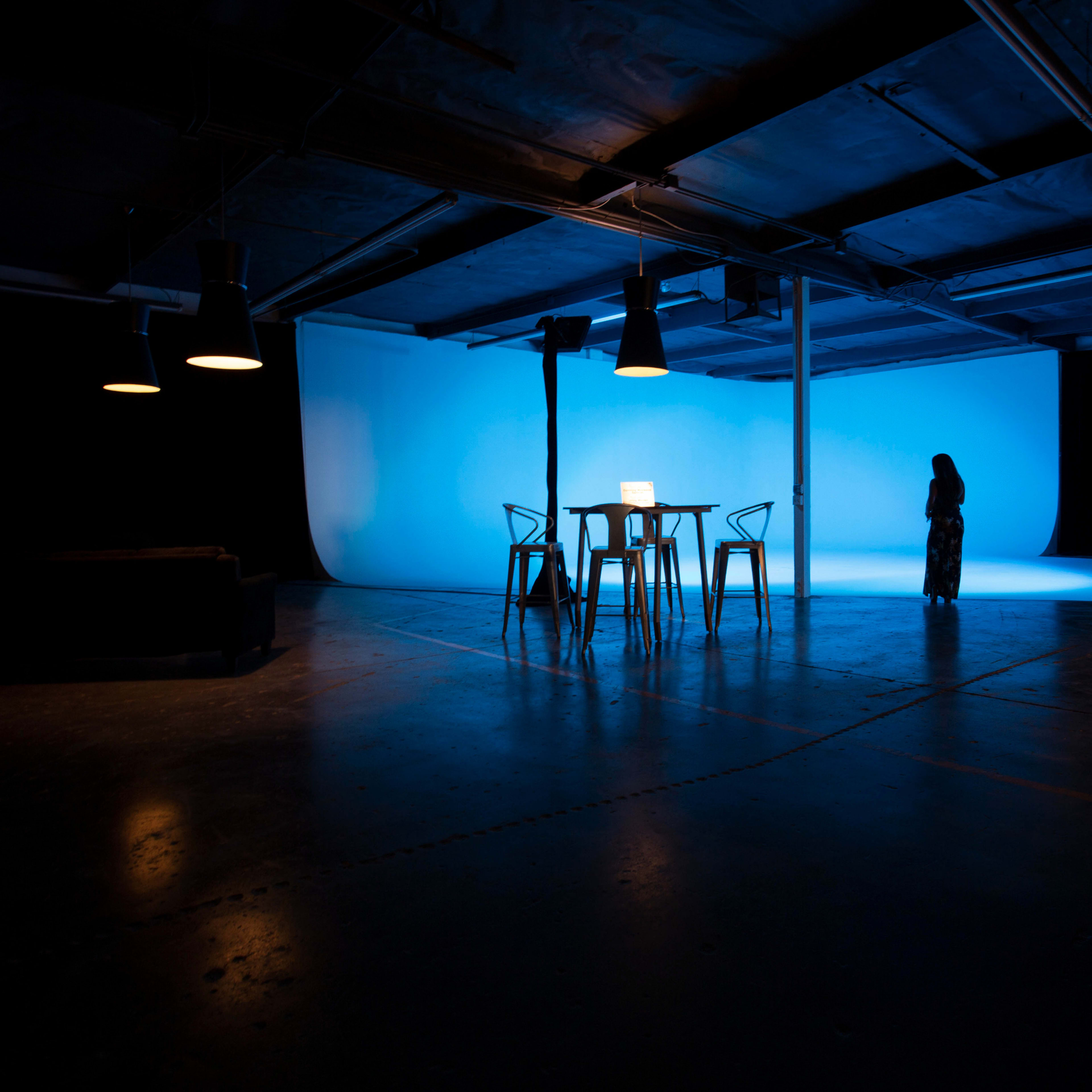 A dimly lit studio features a blue backdrop, a table with a laptop, and a solitary figure standing nearby.