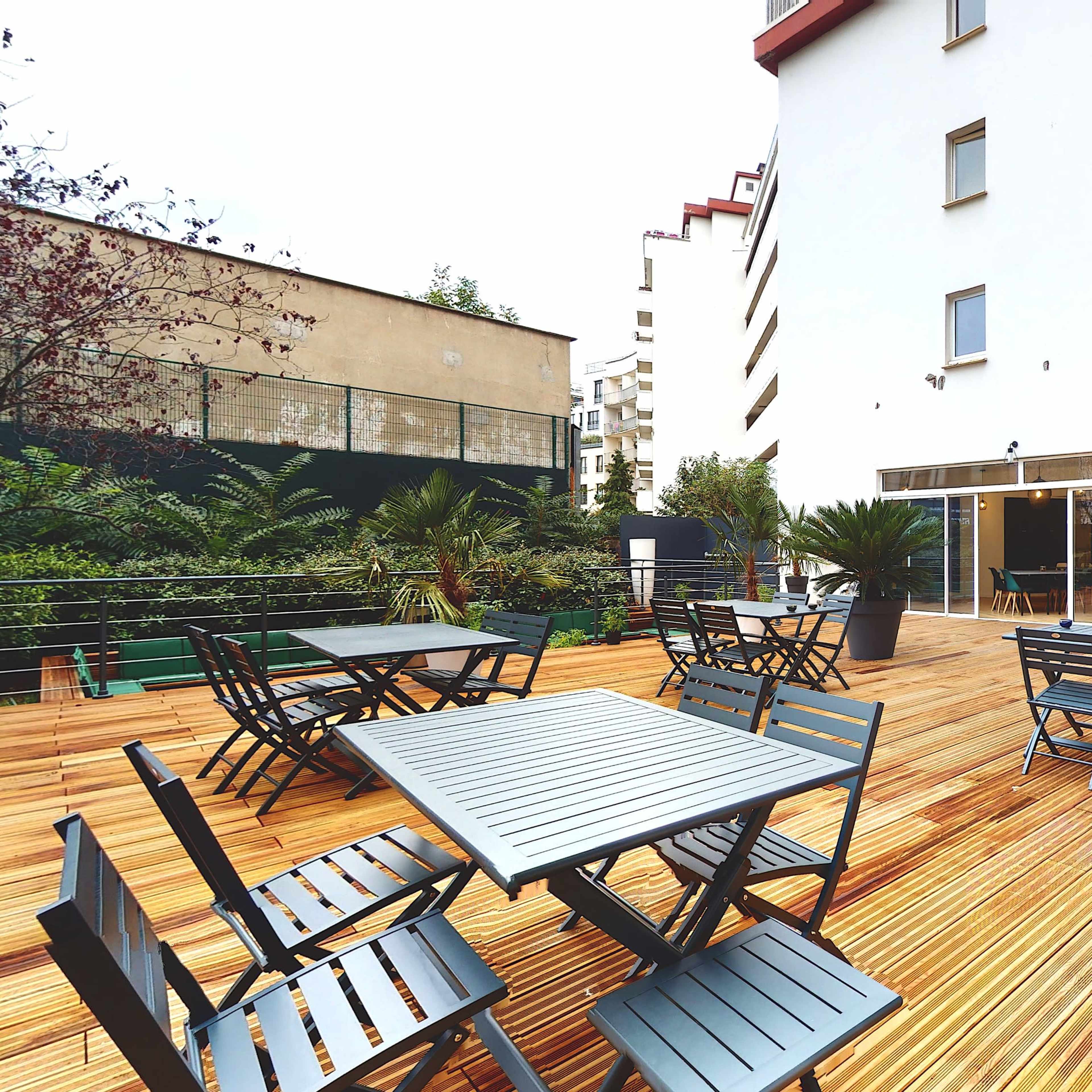 A wooden deck area with several black metal tables and chairs, surrounded by plants and adjacent to a building with large glass doors.