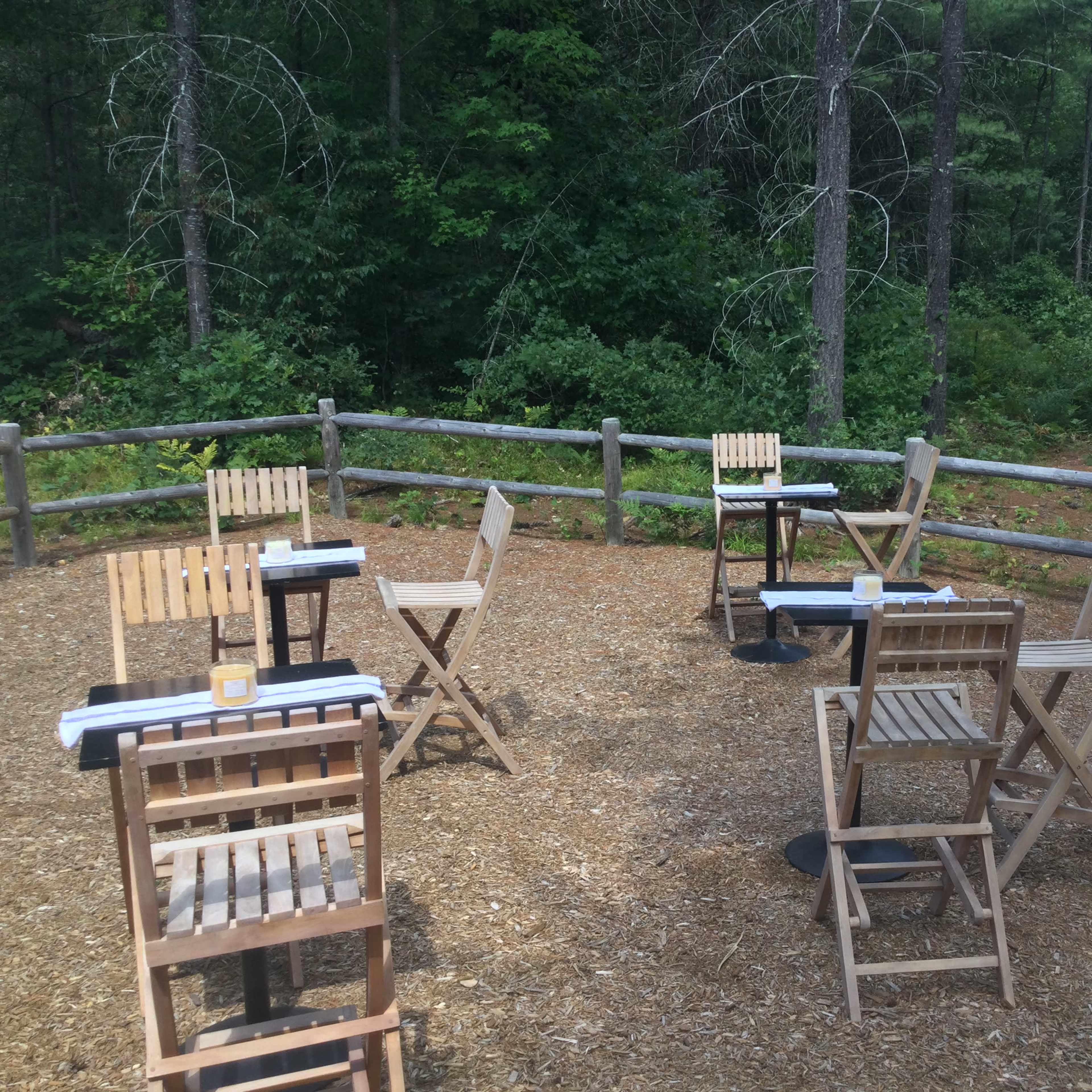 A small outdoor seating area with several wooden chairs and tables arranged on a gravel surface, surrounded by trees and featuring a red umbrella on a nearby table.