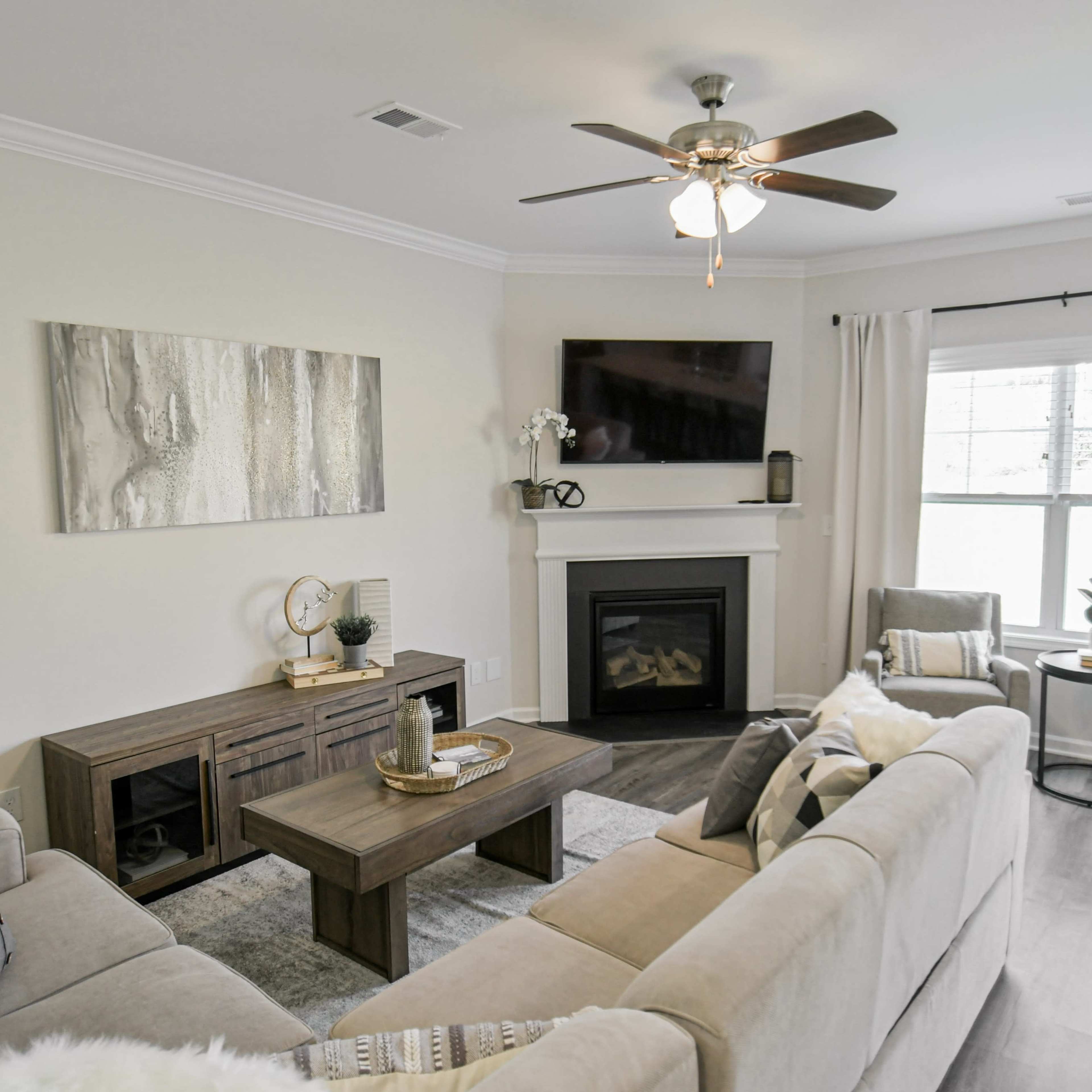 A modern living room features a beige sectional sofa, a wooden coffee table, and a fireplace, with large windows covered by light curtains.