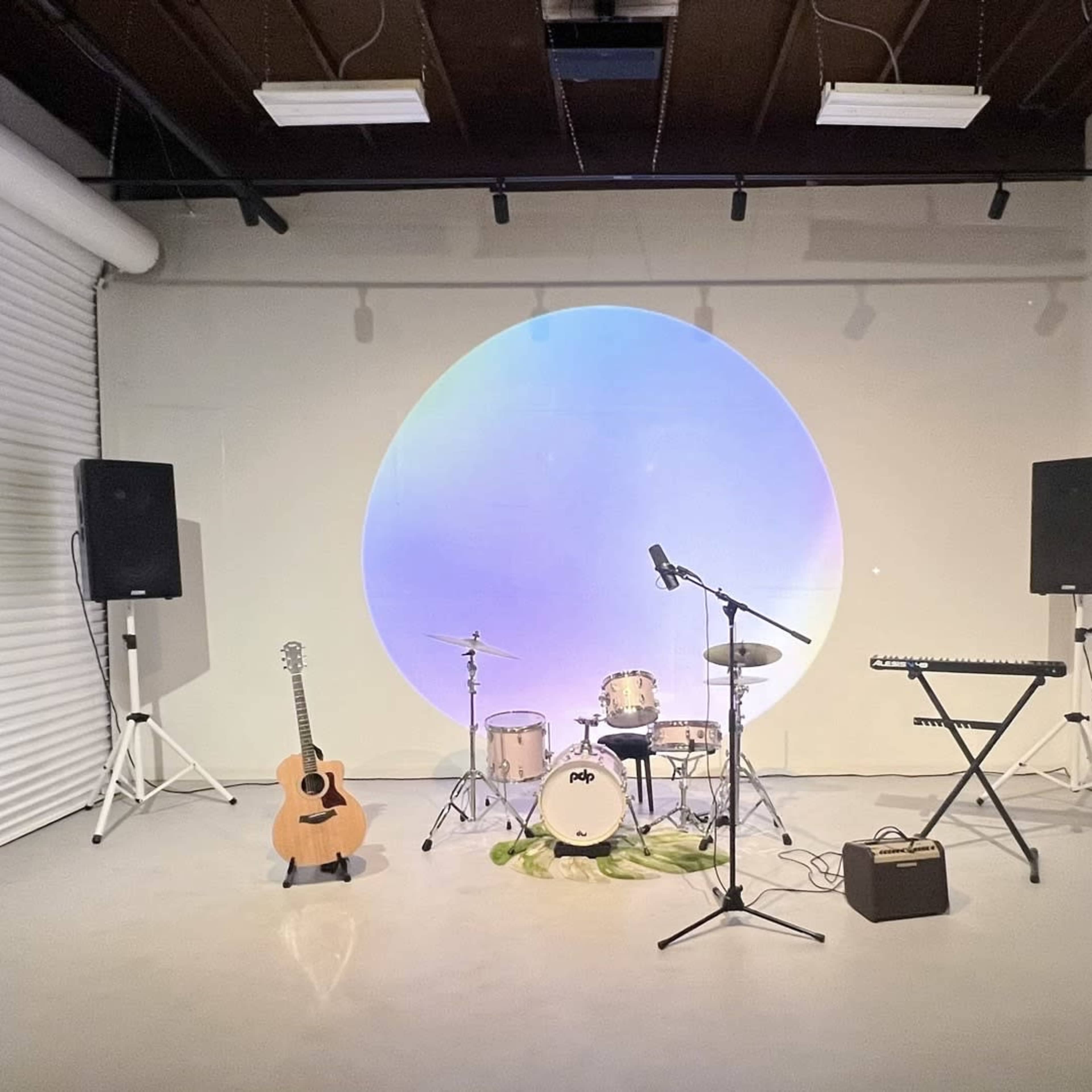A minimalist performance space featuring a drum set, guitar, keyboard, and two speakers, with a large circular projection on the wall.