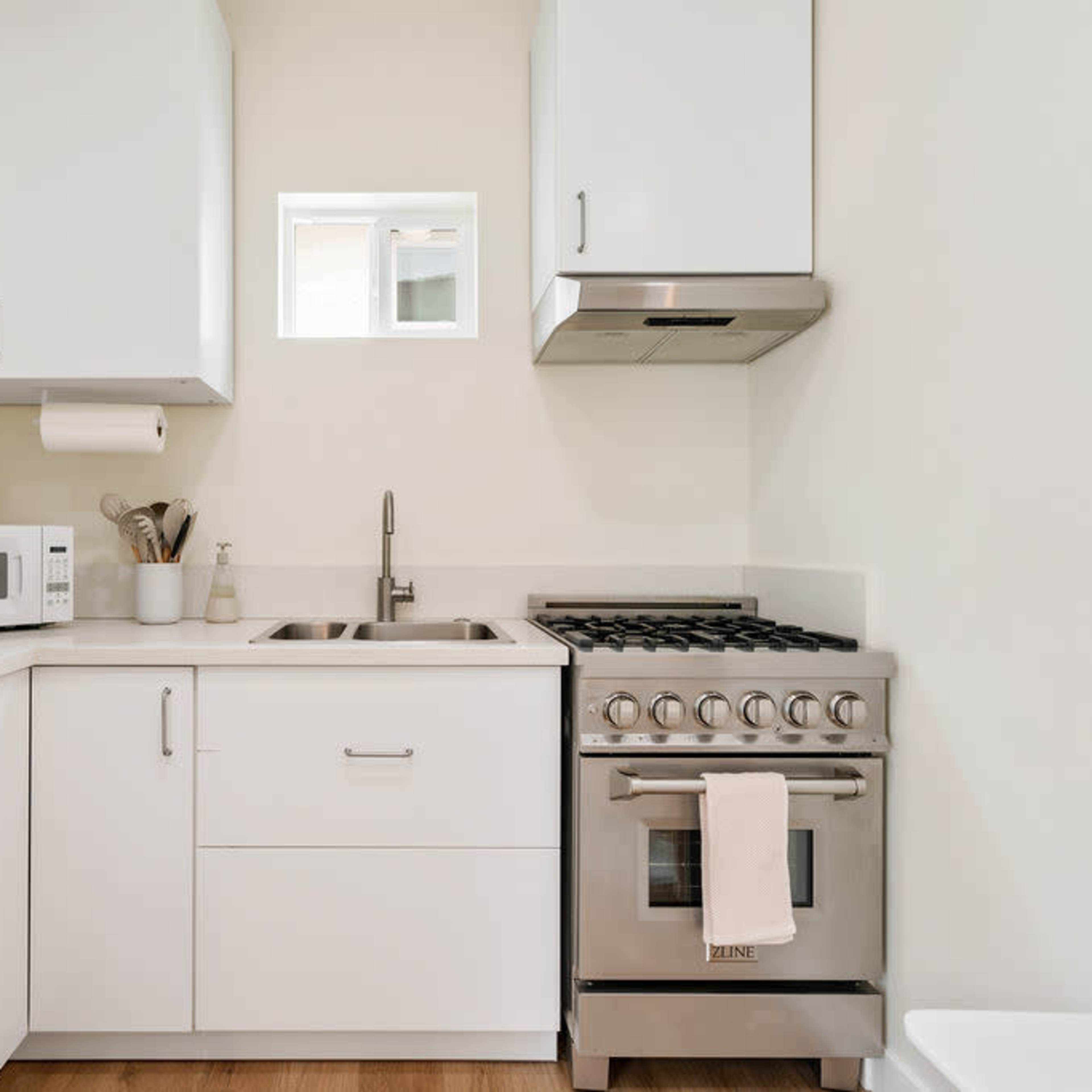A compact kitchen features white cabinetry, a stainless steel gas stove, a microwave, and a small sink with a window above it.