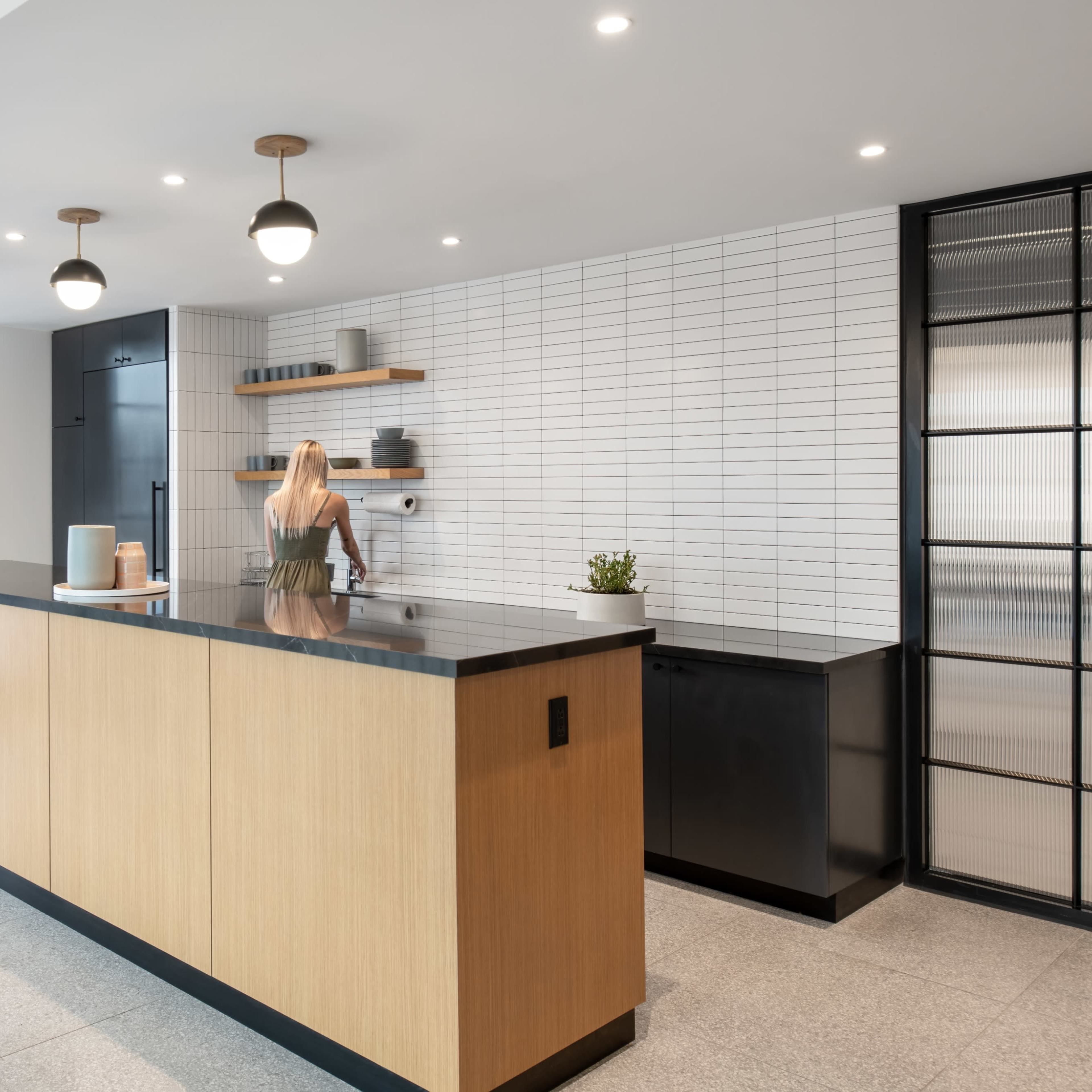 A modern kitchen features a long wooden island, sleek black cabinetry, and bright overhead lighting, with two people present in the space.