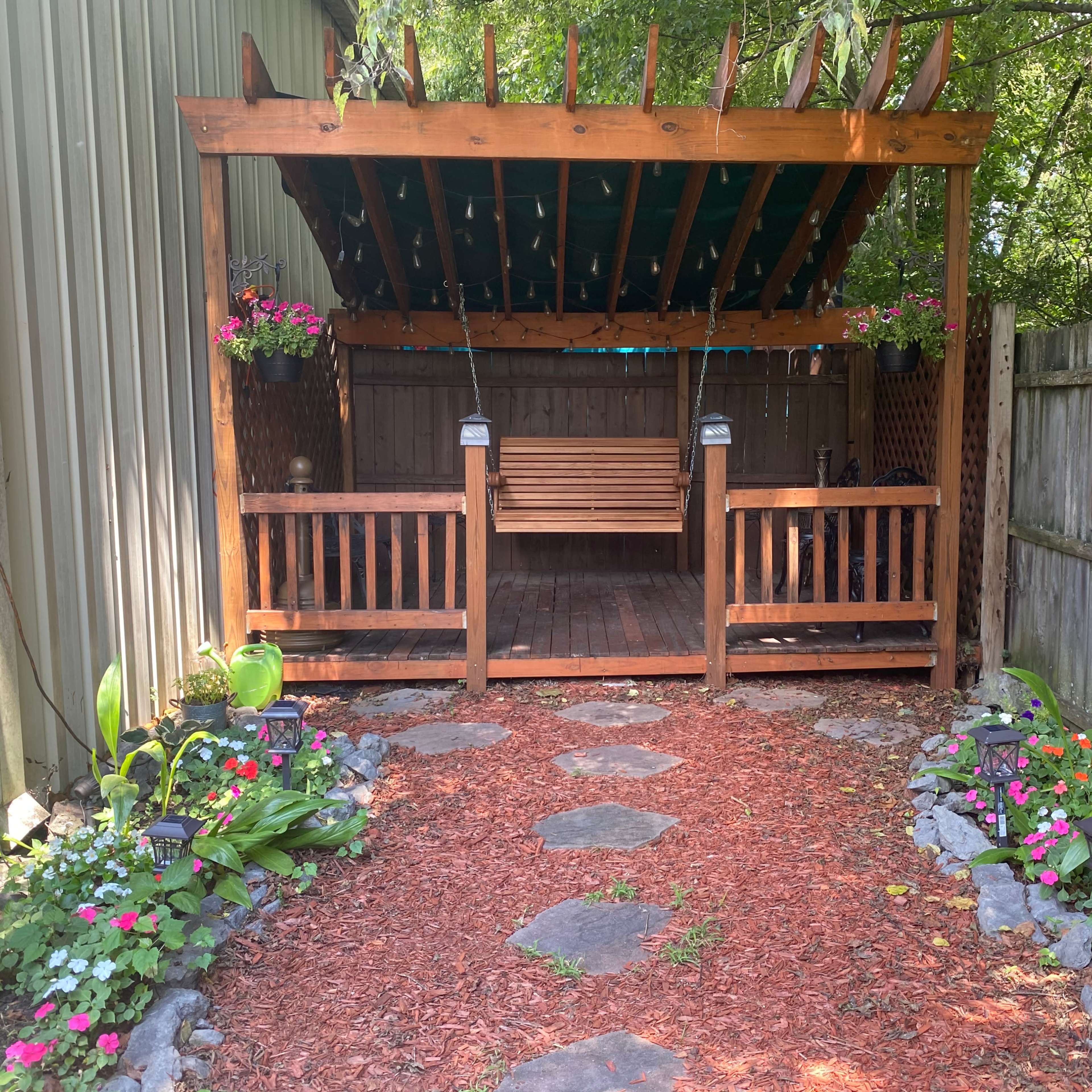 A wooden gazebo with a swinging bench is surrounded by a stone pathway adorned with flower beds and hanging plants.