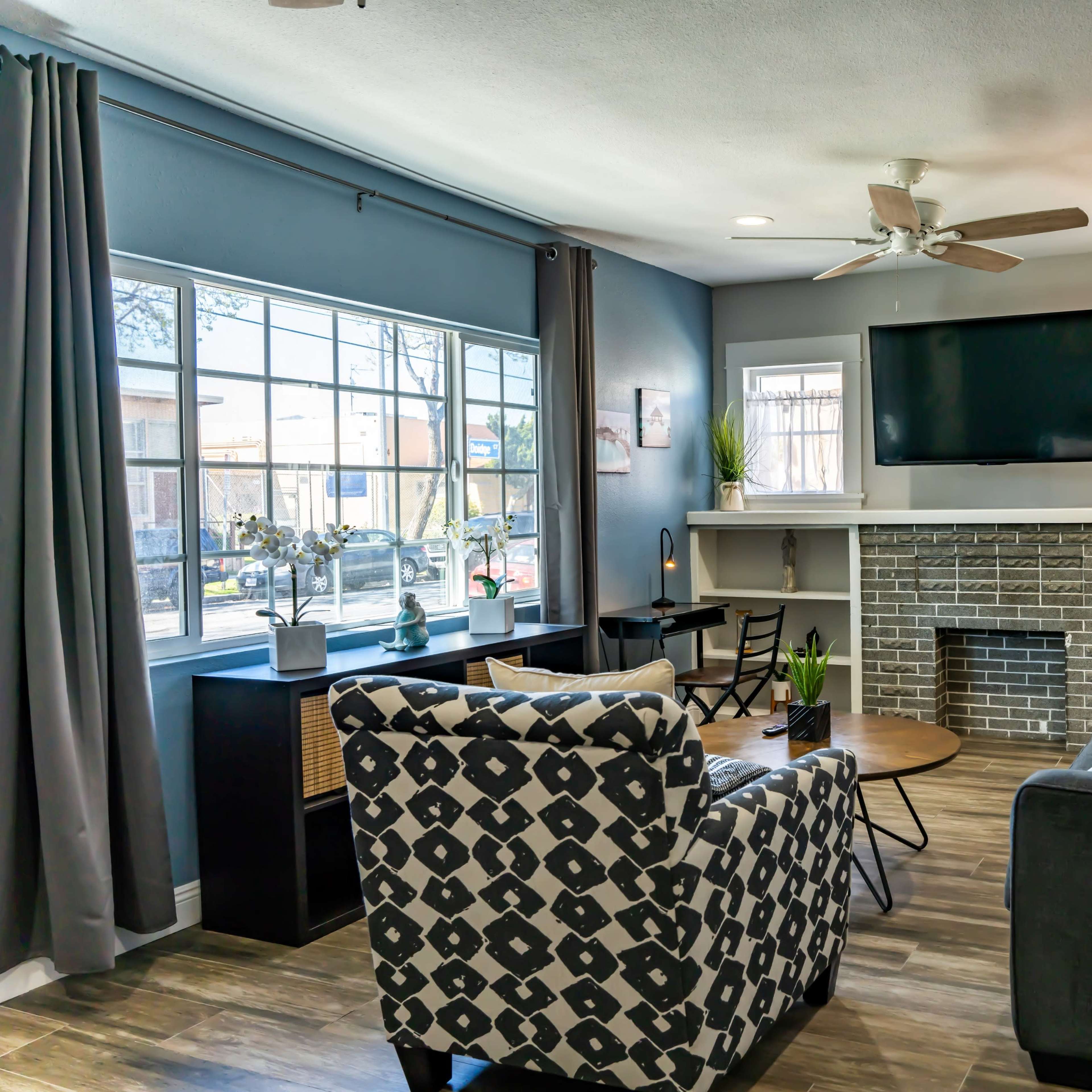 A modern living room with a gray color scheme, featuring a sectional sofa, an armchair, a coffee table, a flat-screen TV mounted above a brick fireplace, and large windows allowing natural light to enter.