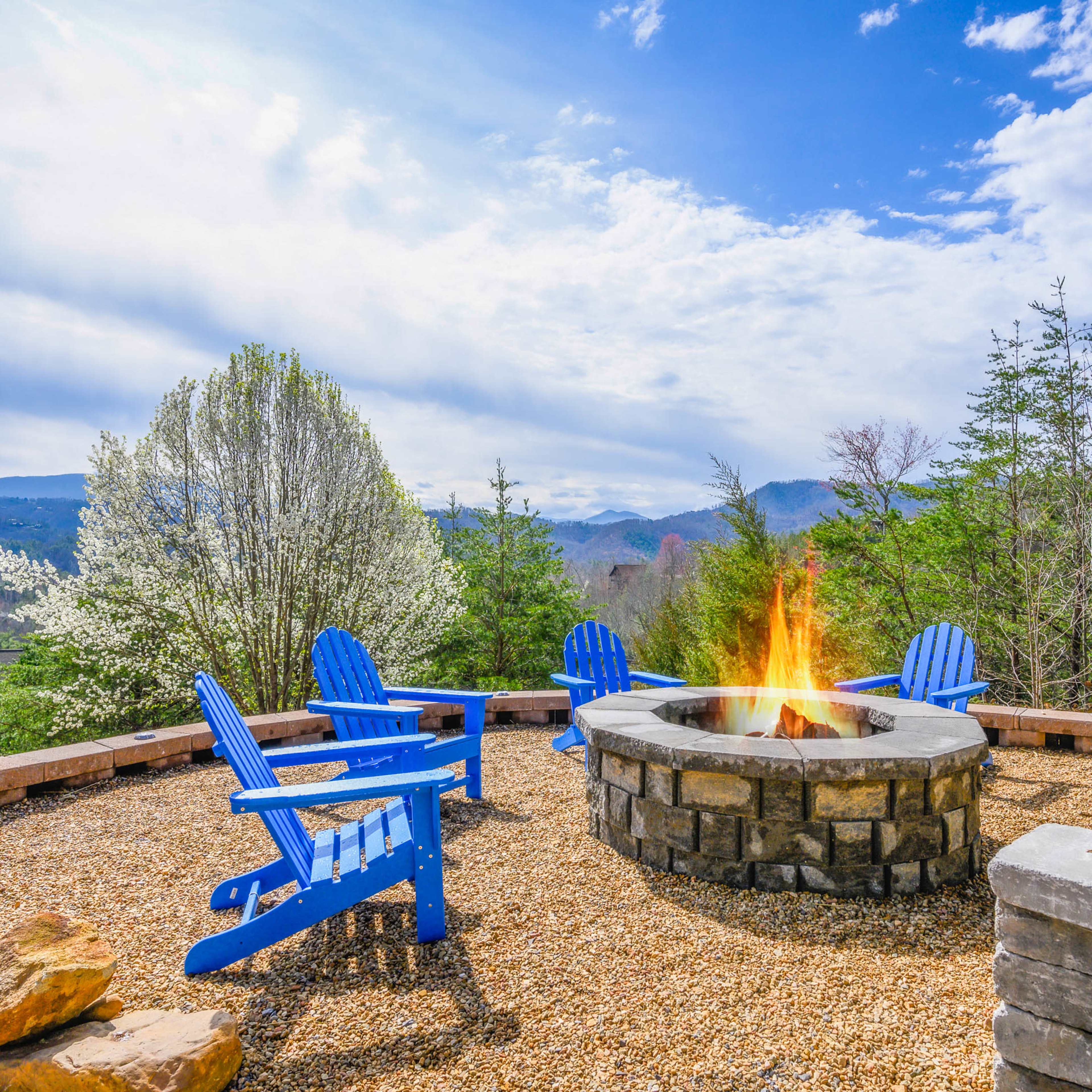 A stone fire pit surrounded by blue Adirondack chairs sits on a gravel patio, with trees and mountains visible in the background under a partly cloudy sky.