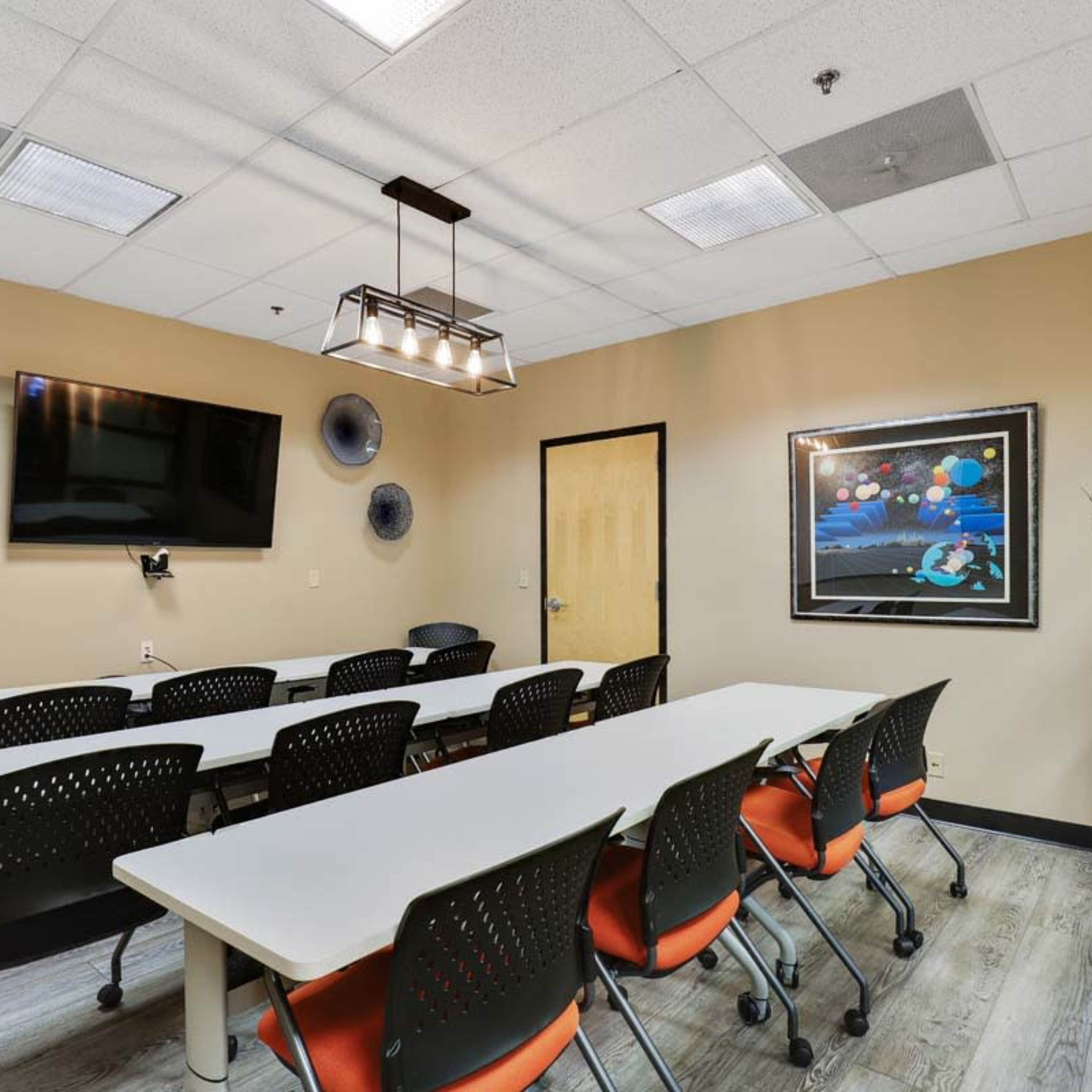 A well-lit meeting room features a long table with rolling chairs, a wall-mounted TV, and musical instruments displayed on the walls.