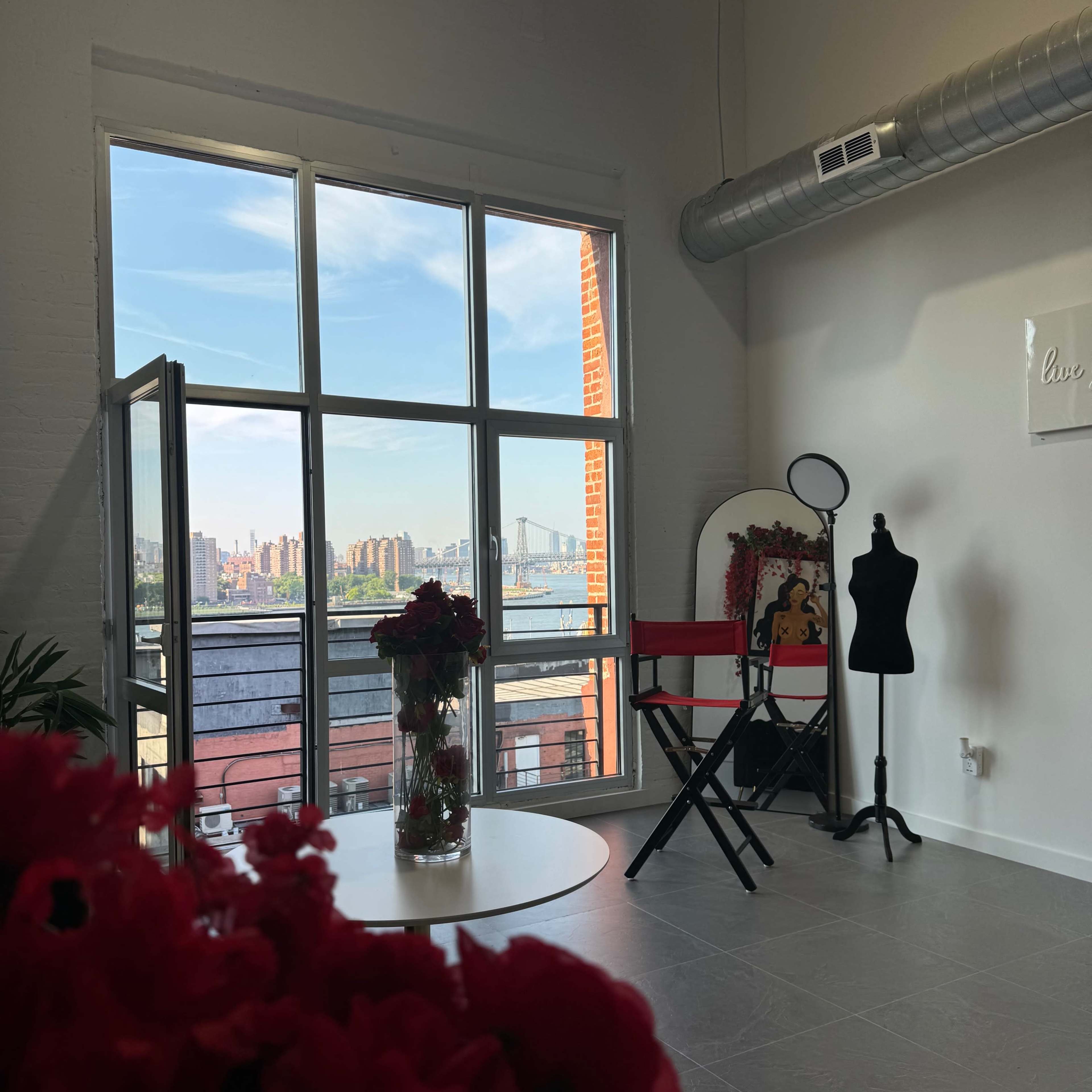 A well-lit studio features a large window overlooking a cityscape, with a table of flowers in the foreground and a mannequin and mirror in the background.
