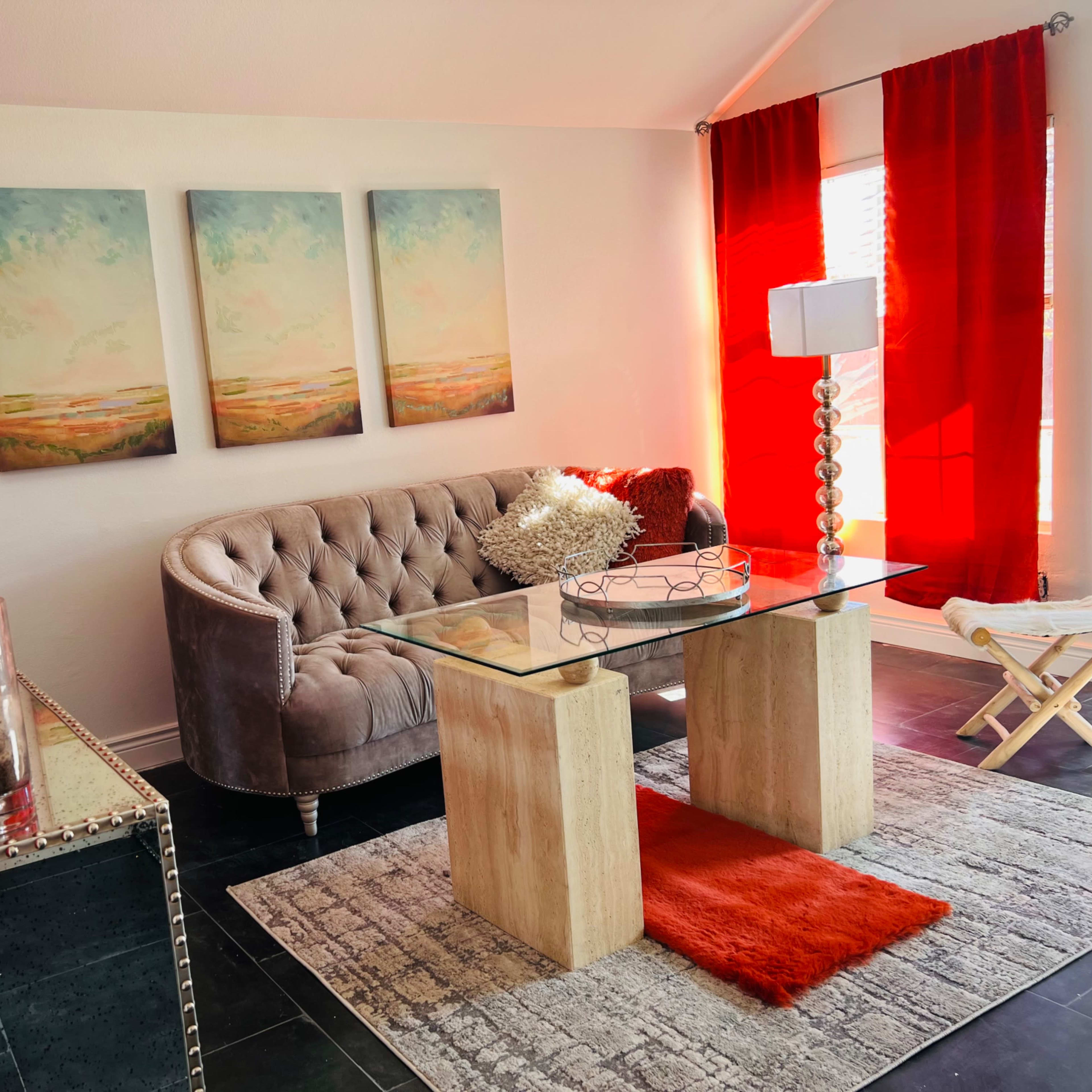 A modern living room with a tufted gray sofa, a glass coffee table, and bright red curtains, complemented by three landscape paintings on the wall.